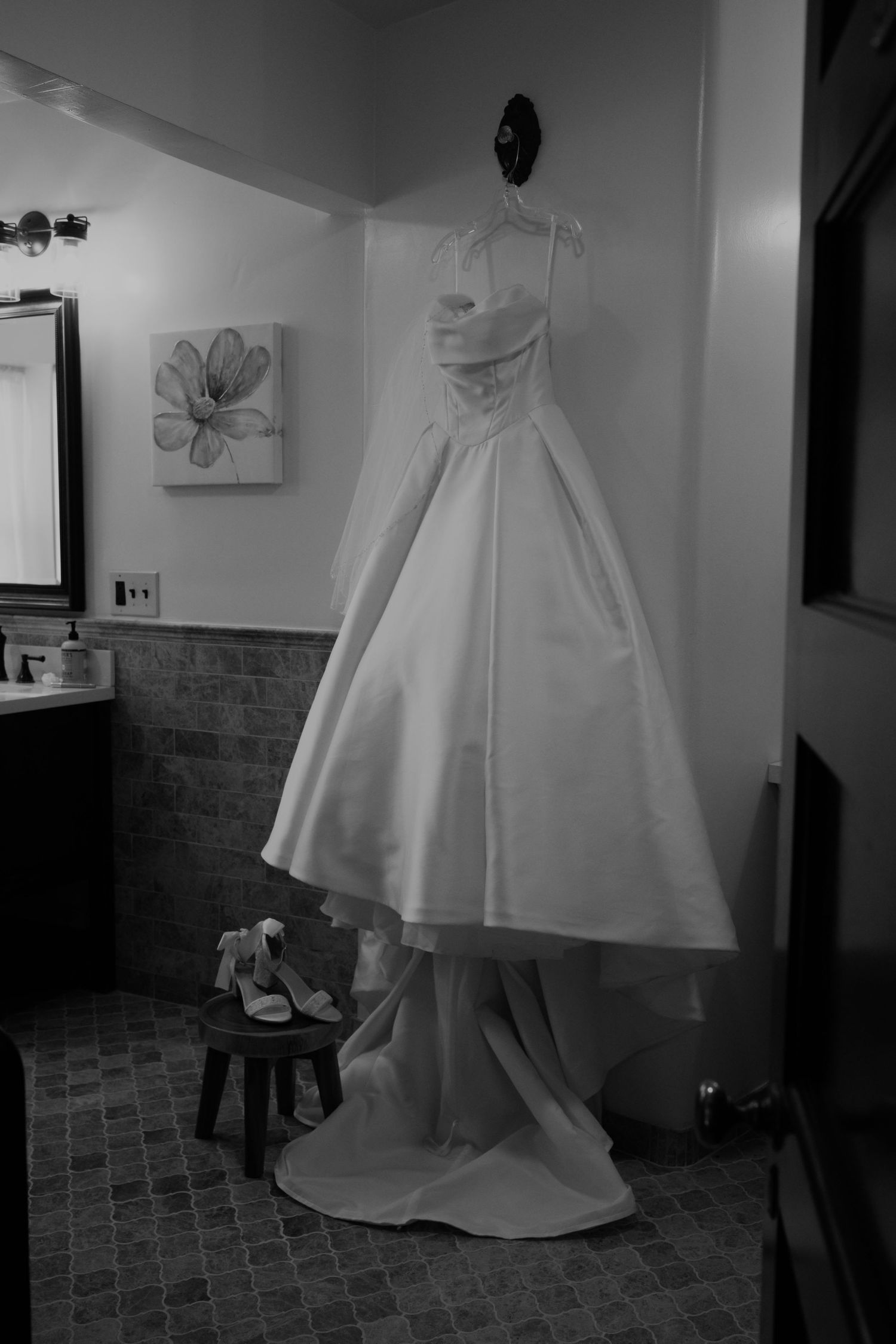 A vintage-style wedding dress hanging in a bathroom with tile walls and floral artwork.