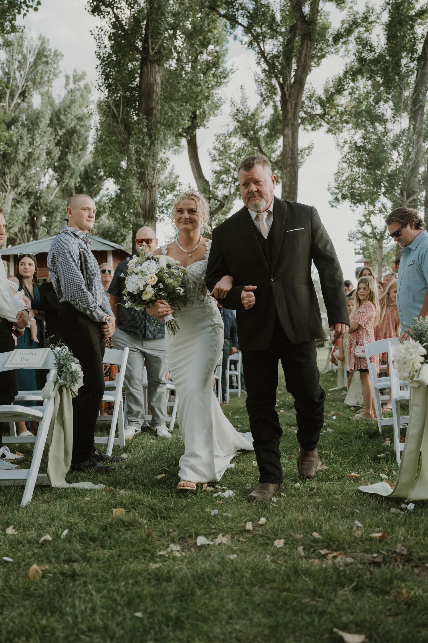 Wedding attendees mingle on a grassy lawn surrounded by mature trees during an outdoor celebration.