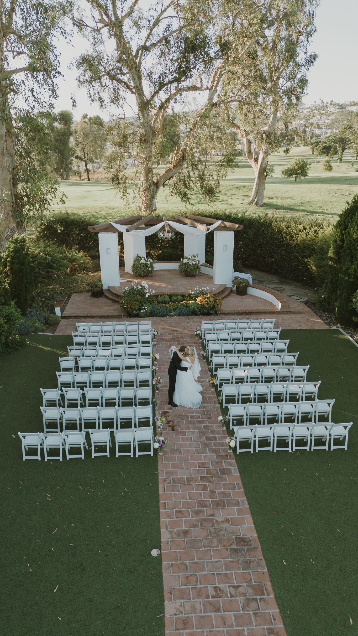 Aerial view of an outdoor wedding ceremony with white chairs, brick aisle, and couple at floral altar surrounded by trees.