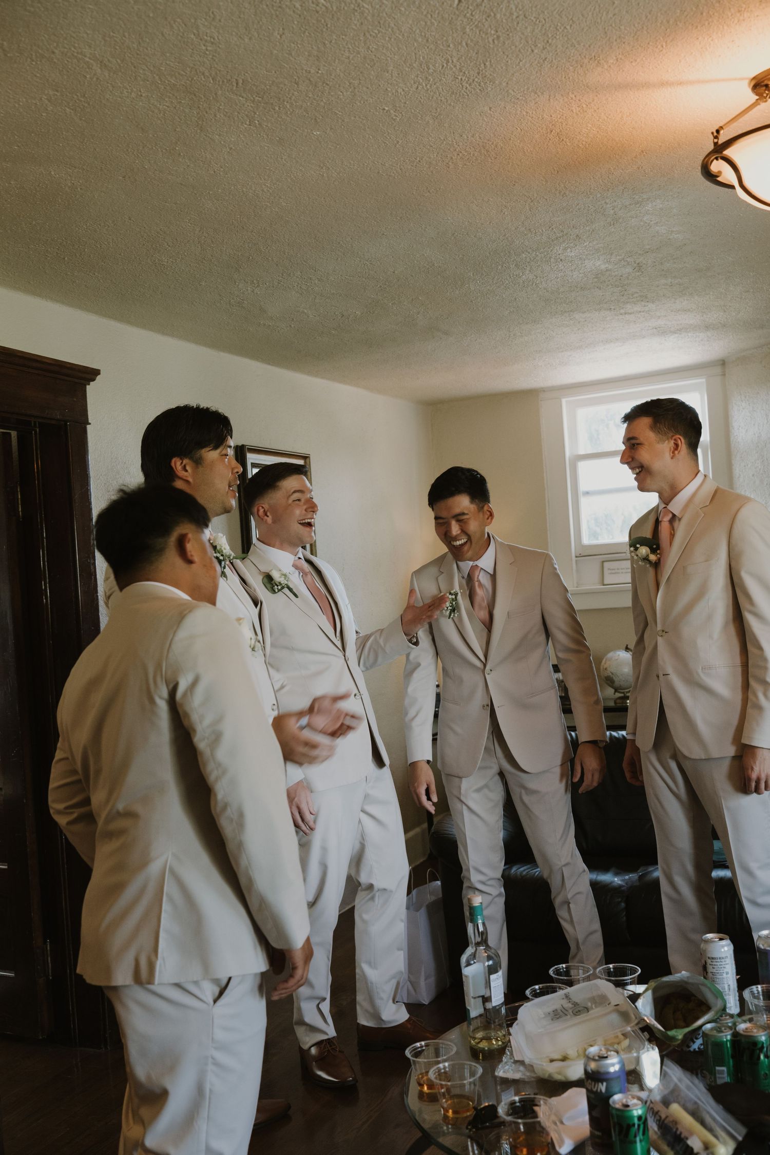 Groomsmen in blush and white suits laughing together in a room before a wedding ceremony.