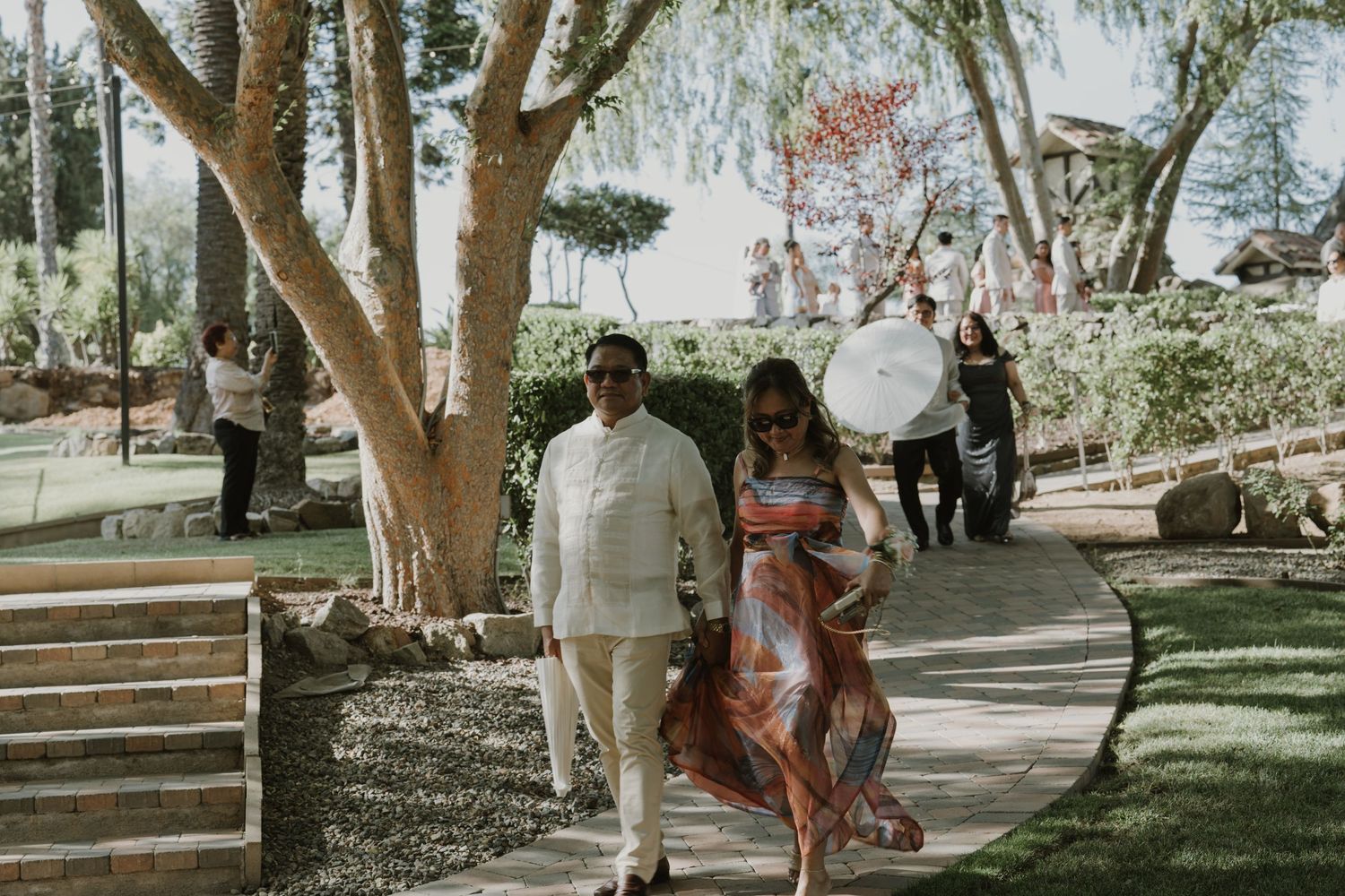 Couple walks along garden path at outdoor wedding venue surrounded by trees.