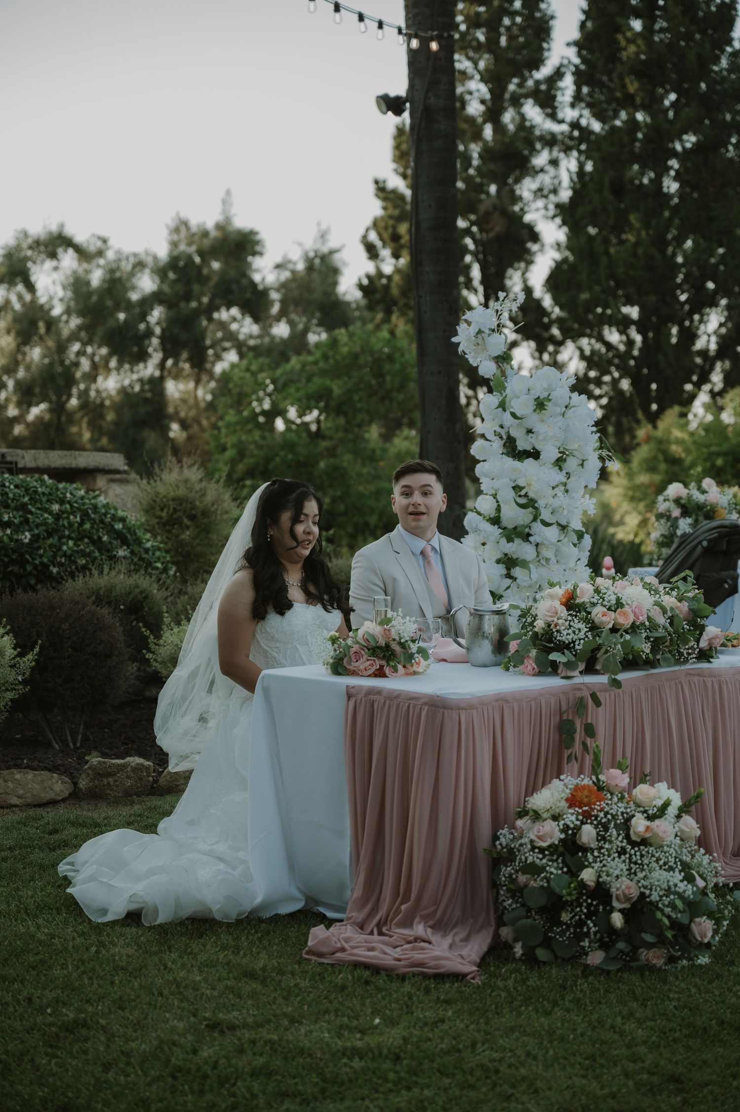 Bride and groom smiling at sweetheart table adorned with pink linens and white floral arch at outdoor wedding reception.