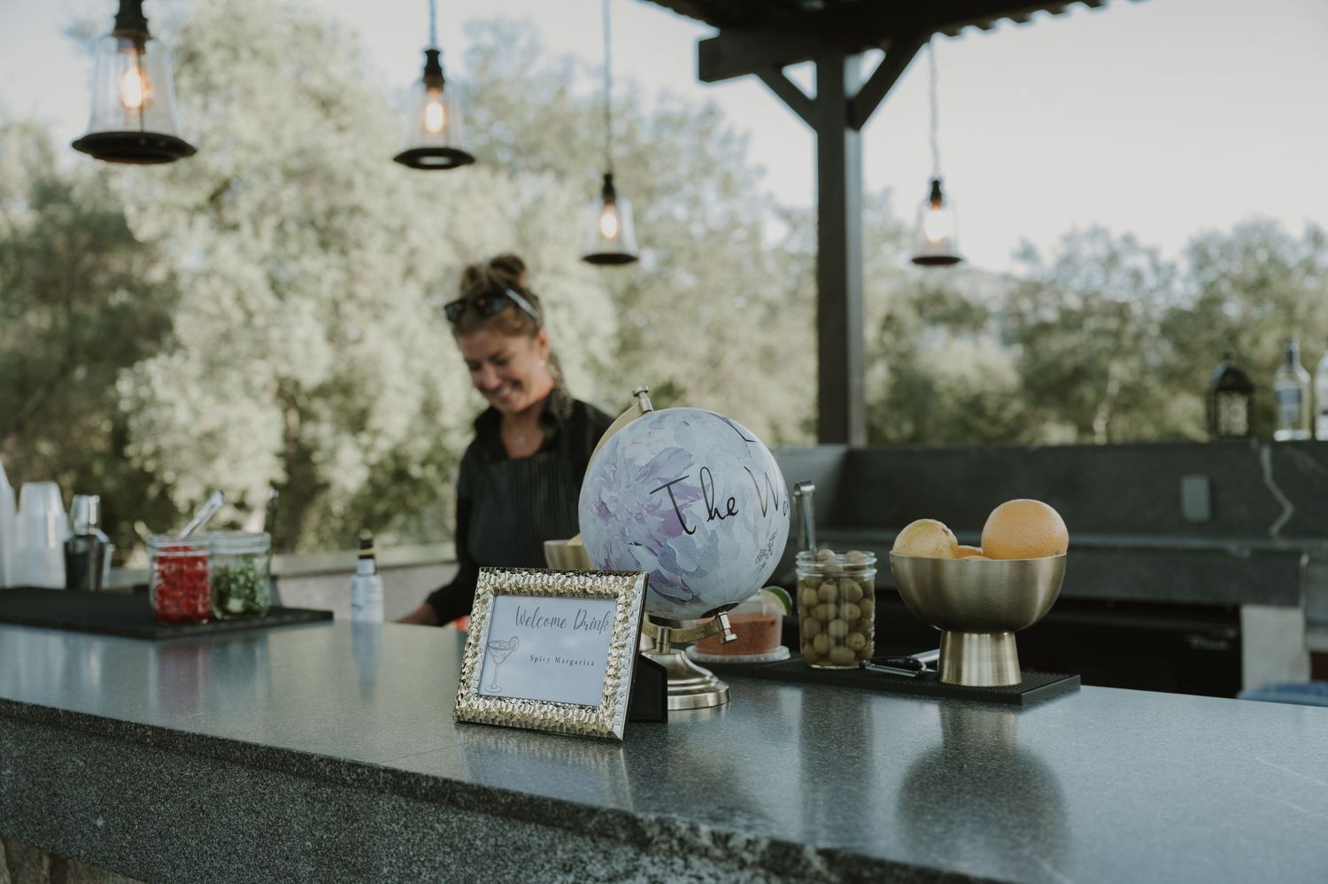 Bartender standing behind an outdoor bar with a decorative globe and framed sign at a rustic event venue.