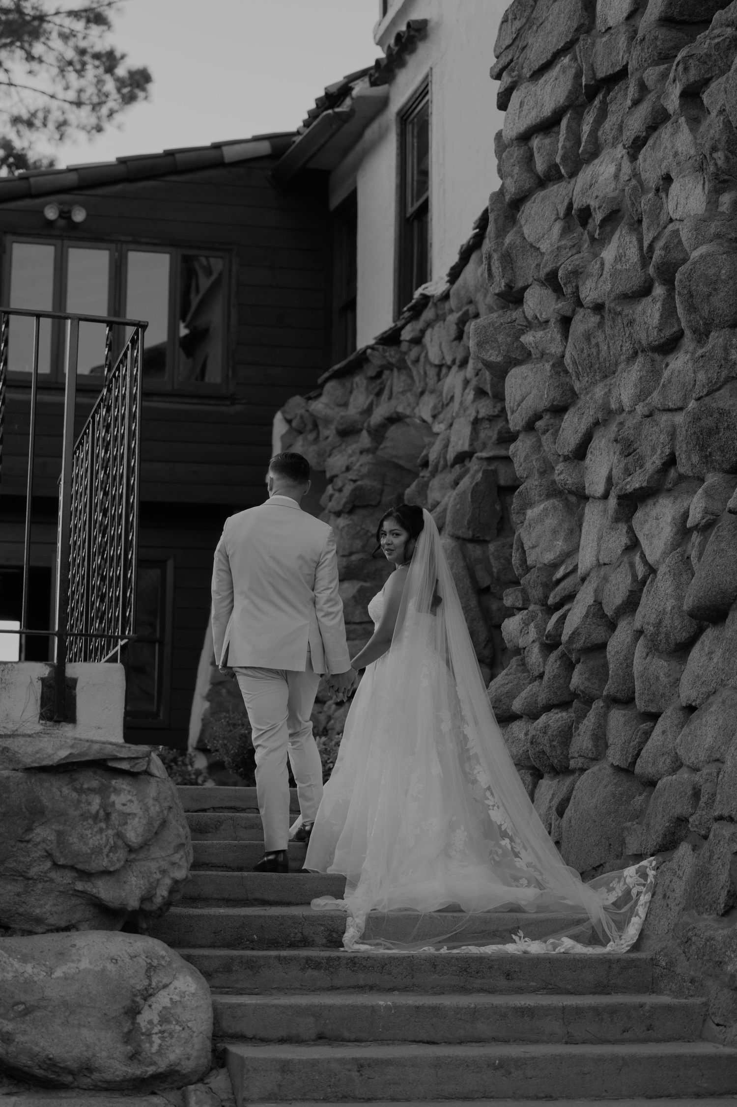 Bride and groom walking up stone steps toward a rustic venue in a black and white wedding photo.