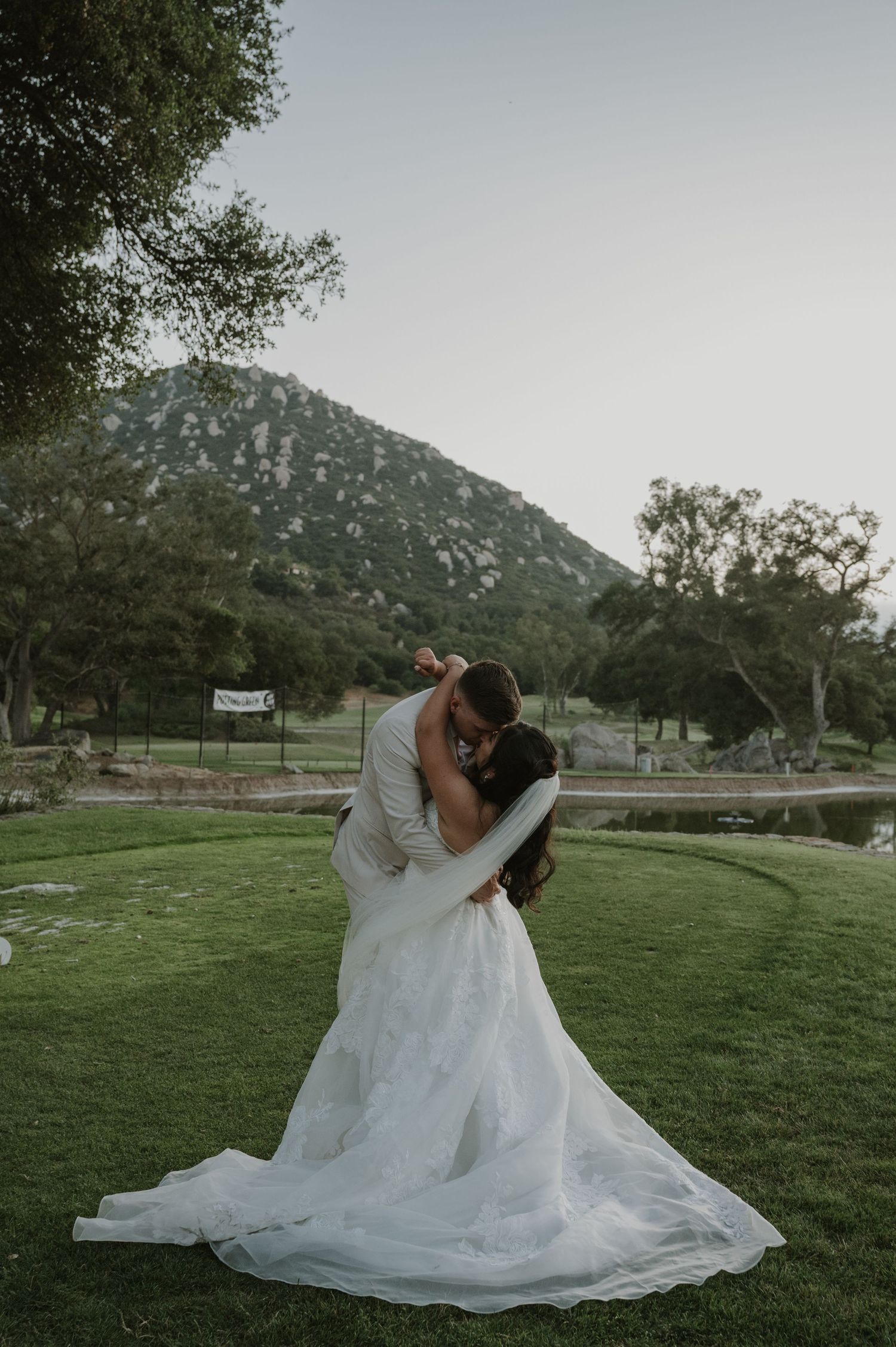 Bride and groom share romantic dip kiss on green lawn with mountain backdrop at outdoor wedding venue.
