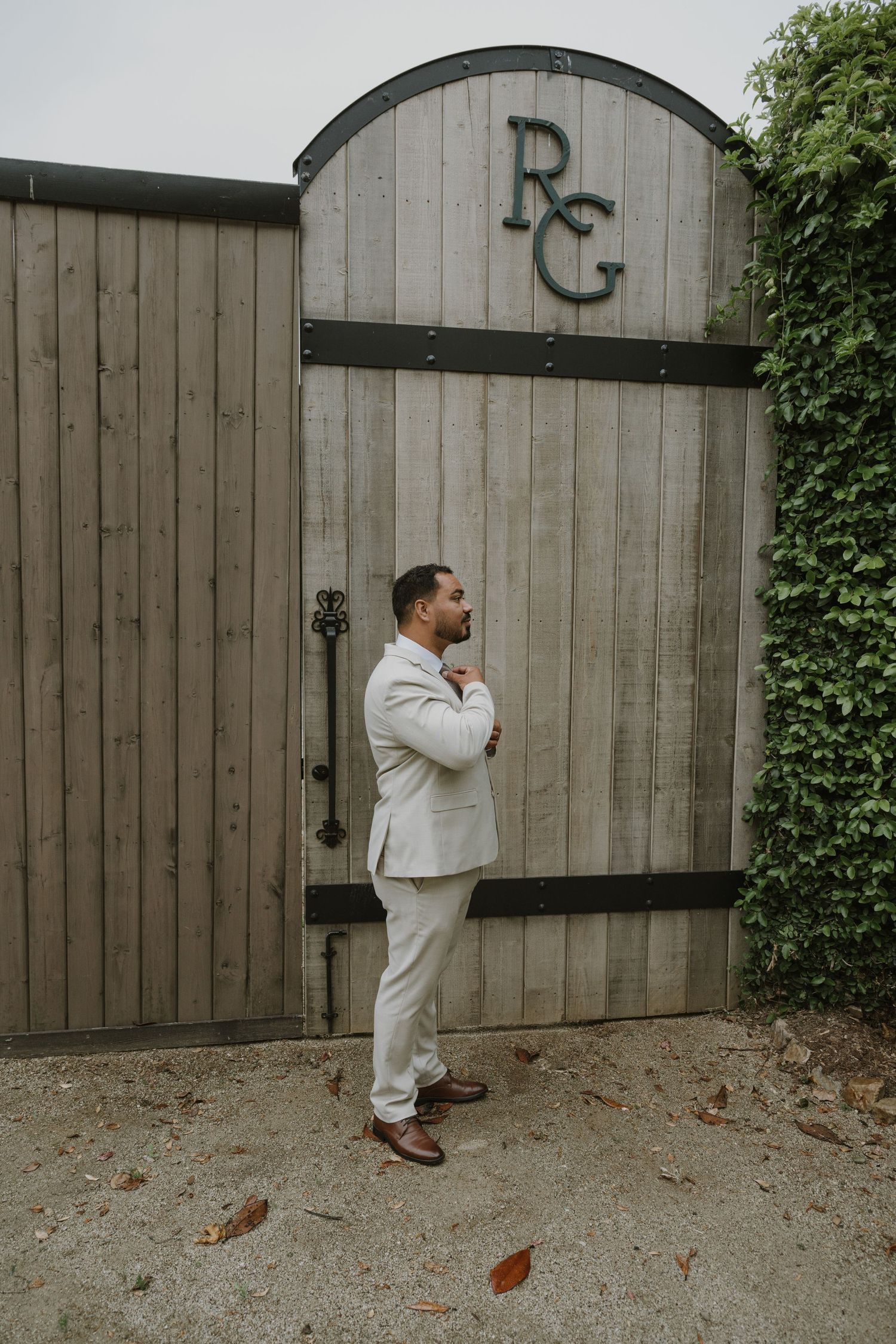 Dapper groom in beige suit stands confidently before rustic wooden gate with RG monogram outdoors.