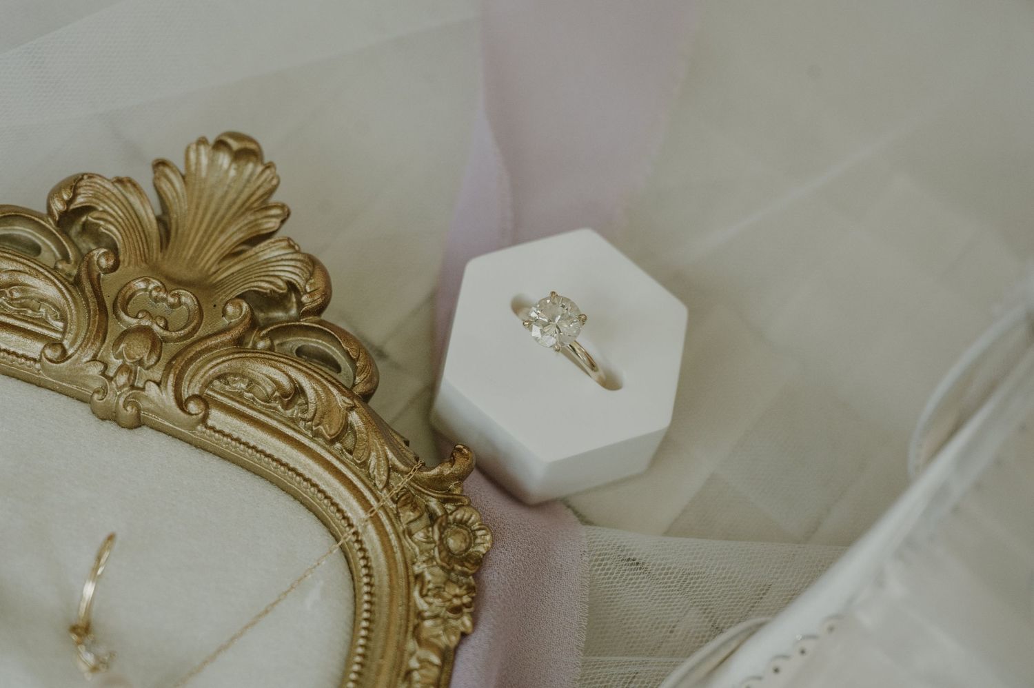Diamond engagement ring in a white hexagonal box beside an ornate gold mirror on soft pink fabric.