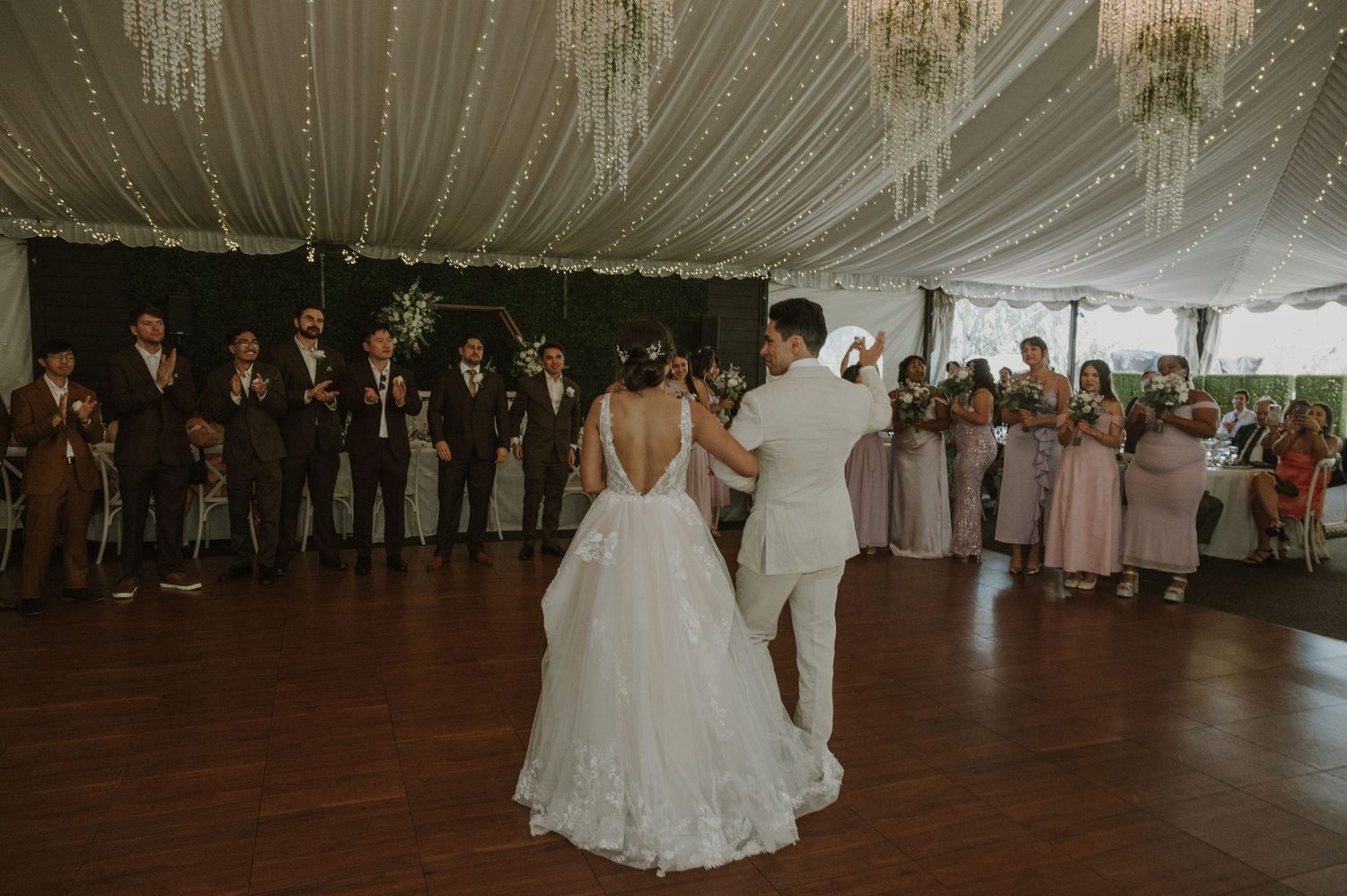 Bride and groom share their first dance as groomsmen and guests watch in a decorated tent reception.