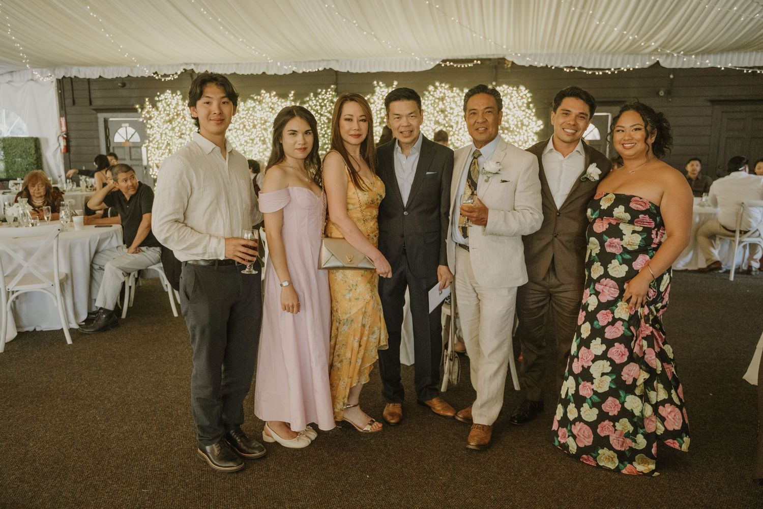 Group of seven wedding guests posing together inside decorated reception tent at celebration.
