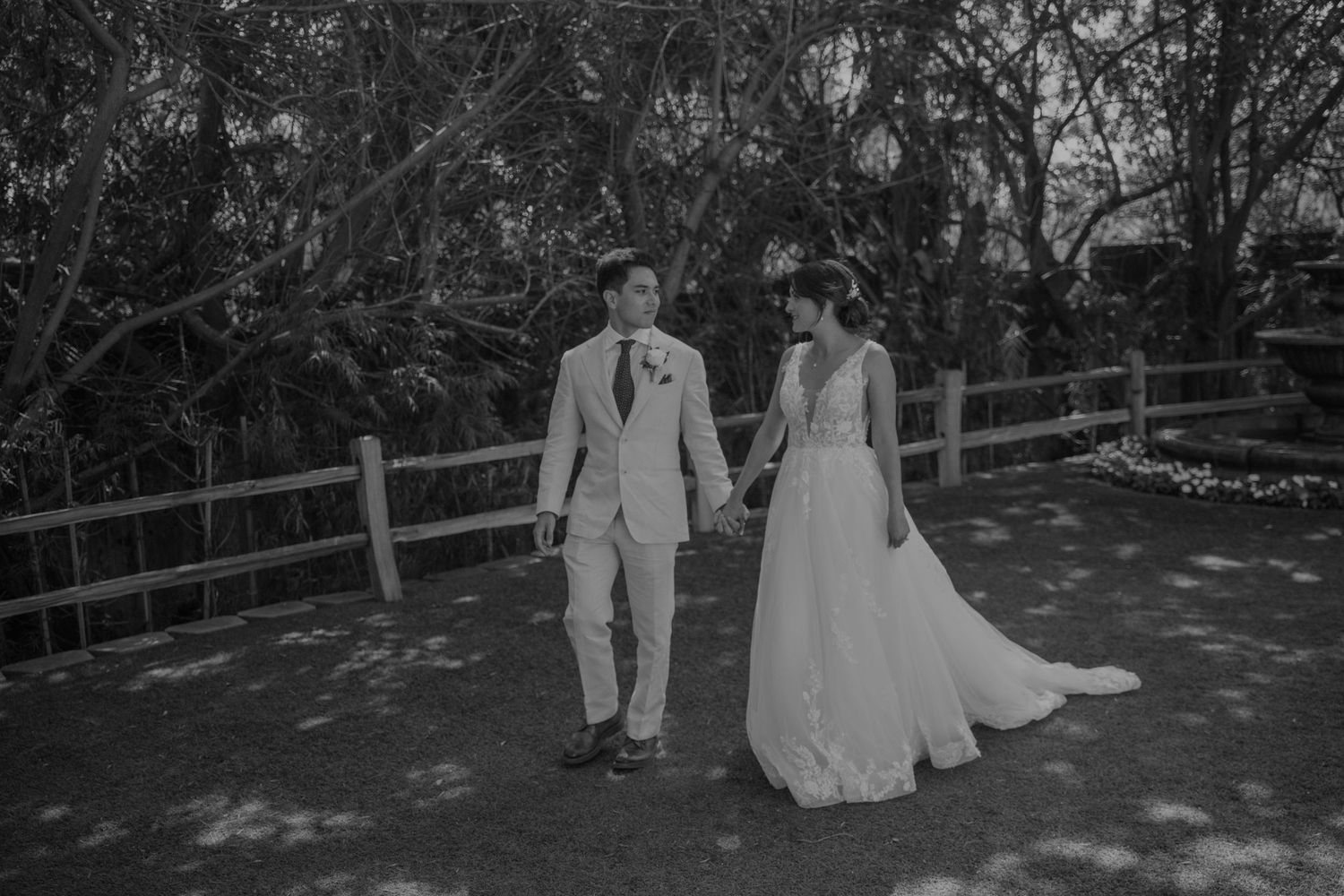 Black and white photo of a bride and groom holding hands on a wooden path surrounded by lush trees.