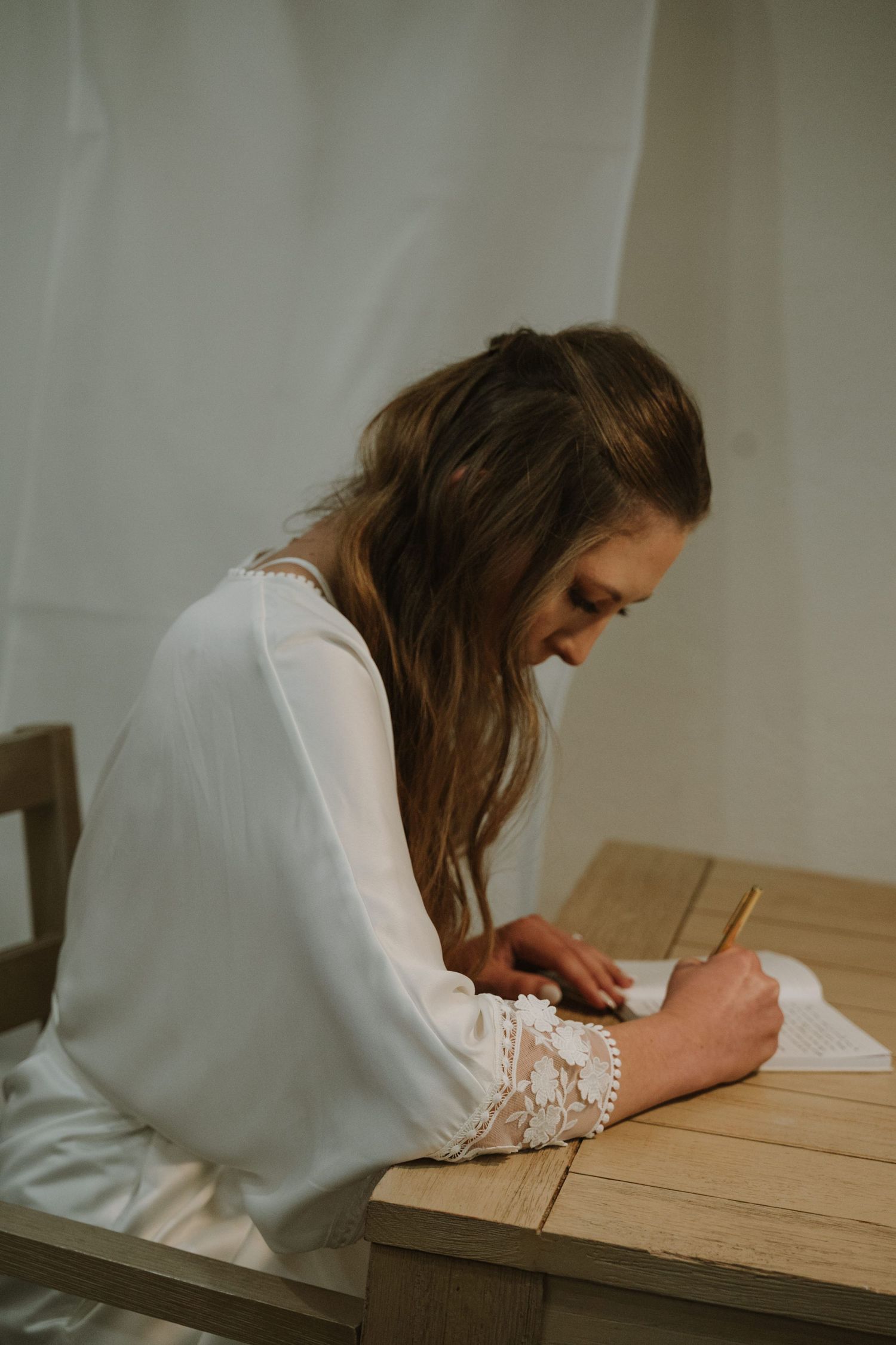Bride in white lace dress writing vows at wooden table with soft natural lighting.