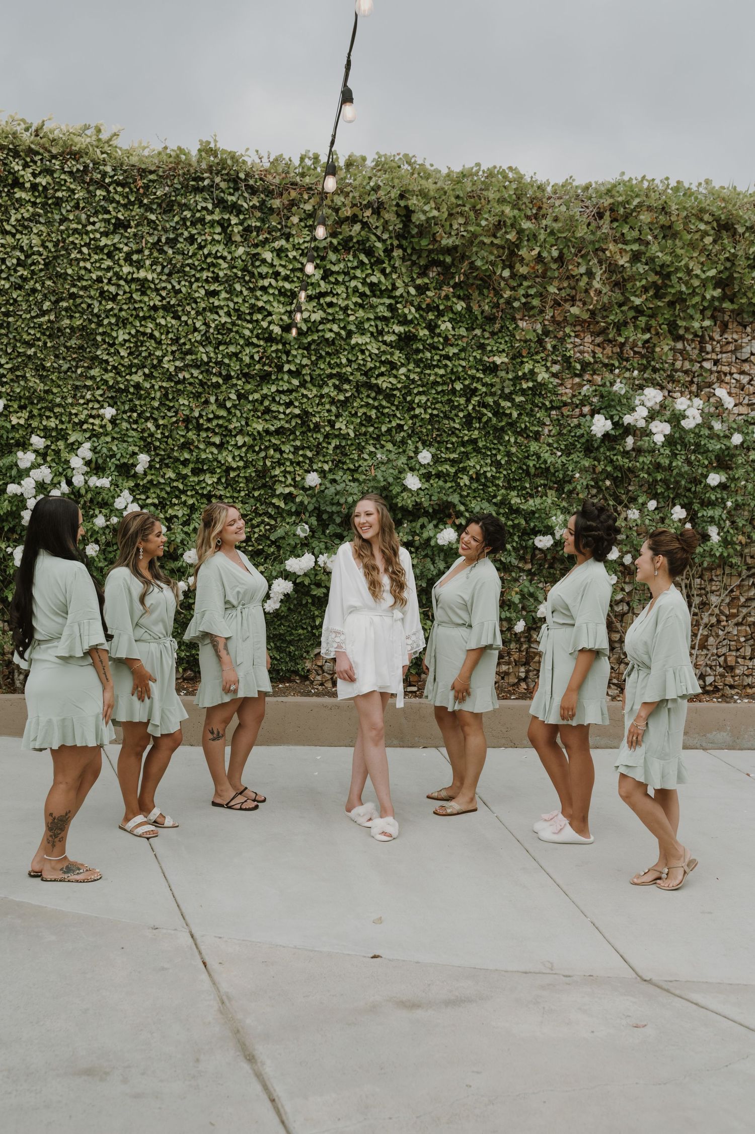 Bride in white robe standing center as bridesmaids in sage green robes gather around her outdoors.