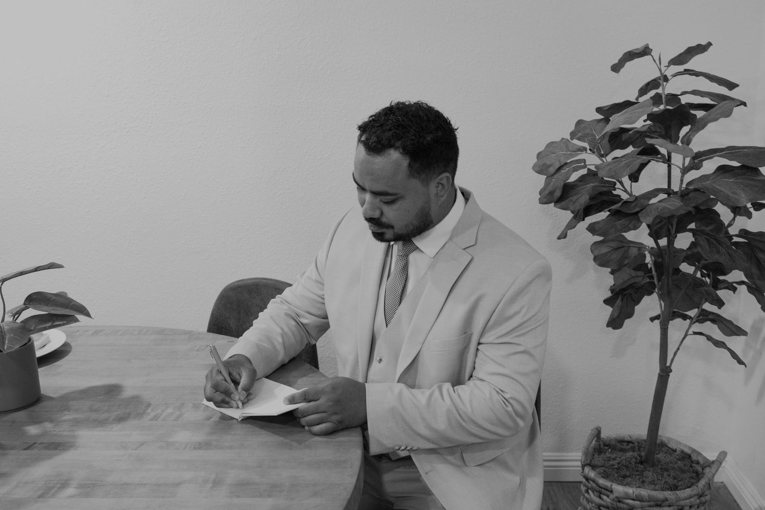 Professional man in suit signing documents at wooden desk beside indoor plant, black and white photo.