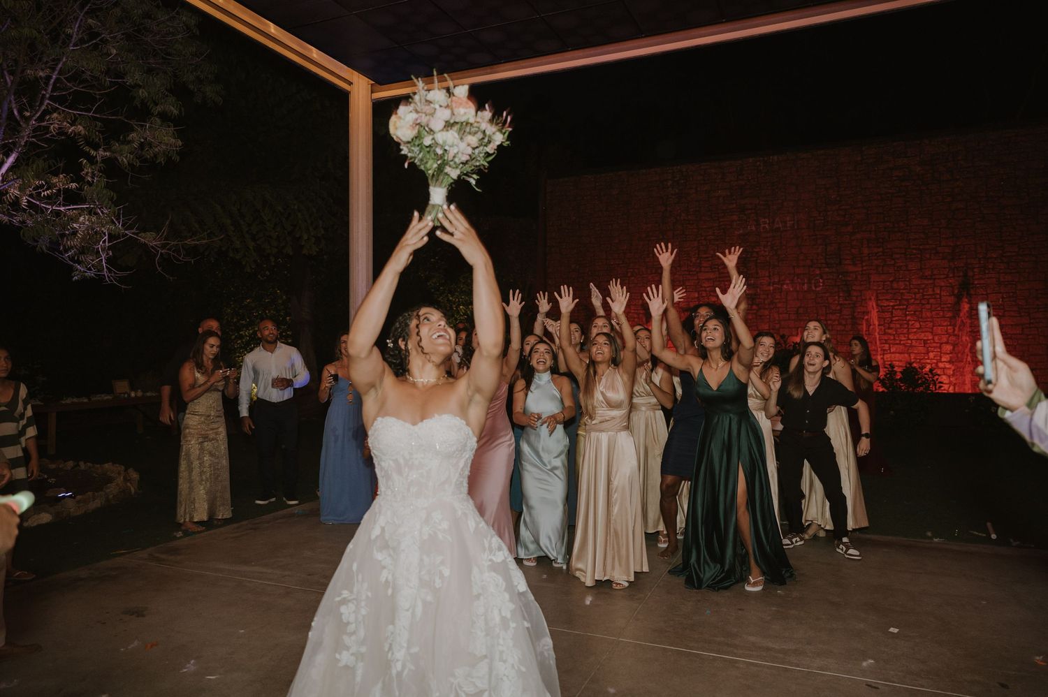 Bride tosses wedding bouquet to excited guests in elegant gowns at outdoor evening reception.