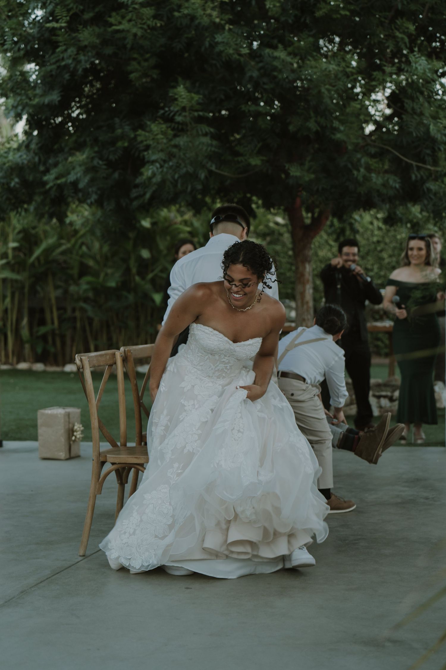 Bride in a strapless white ball gown laughing and dancing with guests at an outdoor wedding reception.