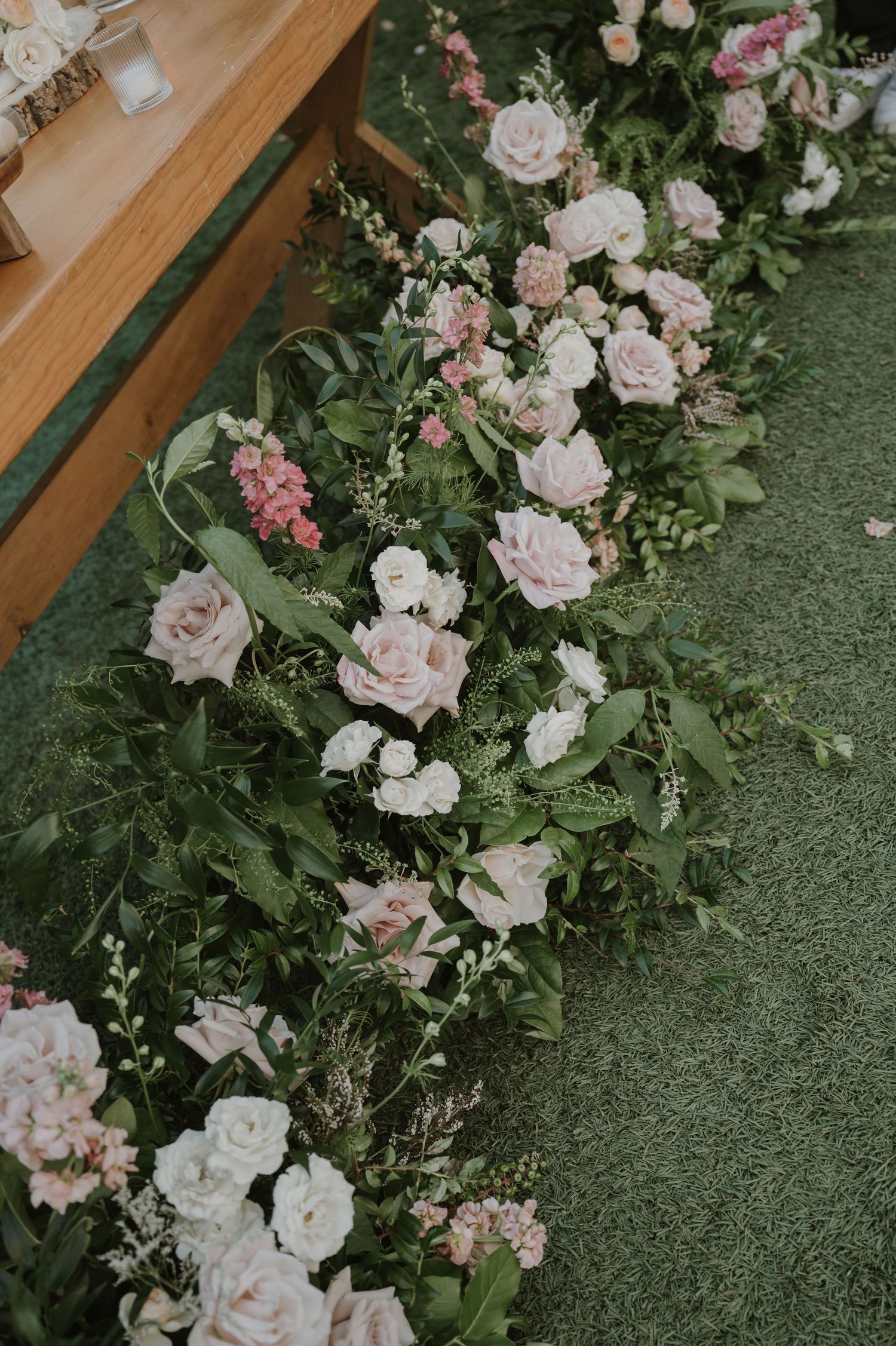 Lush floral arrangement of blush pink and white roses with greenery lining a wedding aisle on green carpet.