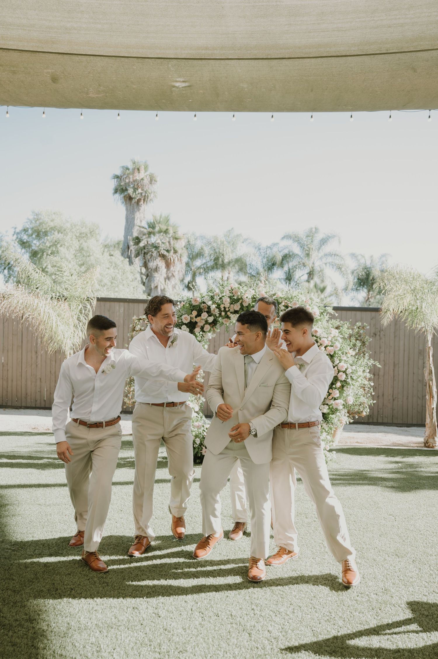 Groom and groomsmen laugh together in neutral suits before a floral arch at an outdoor wedding venue.