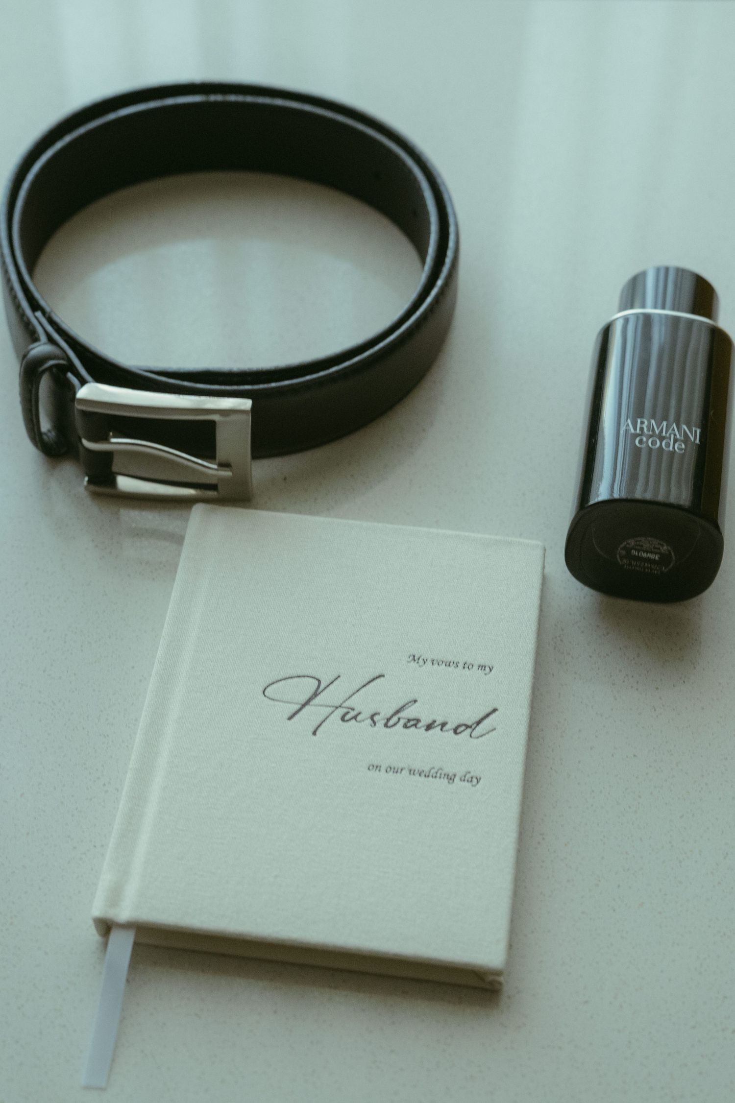 Wedding day gifts for husband including a leather belt, Armani Code cologne, and a heartfelt journal on a white surface.