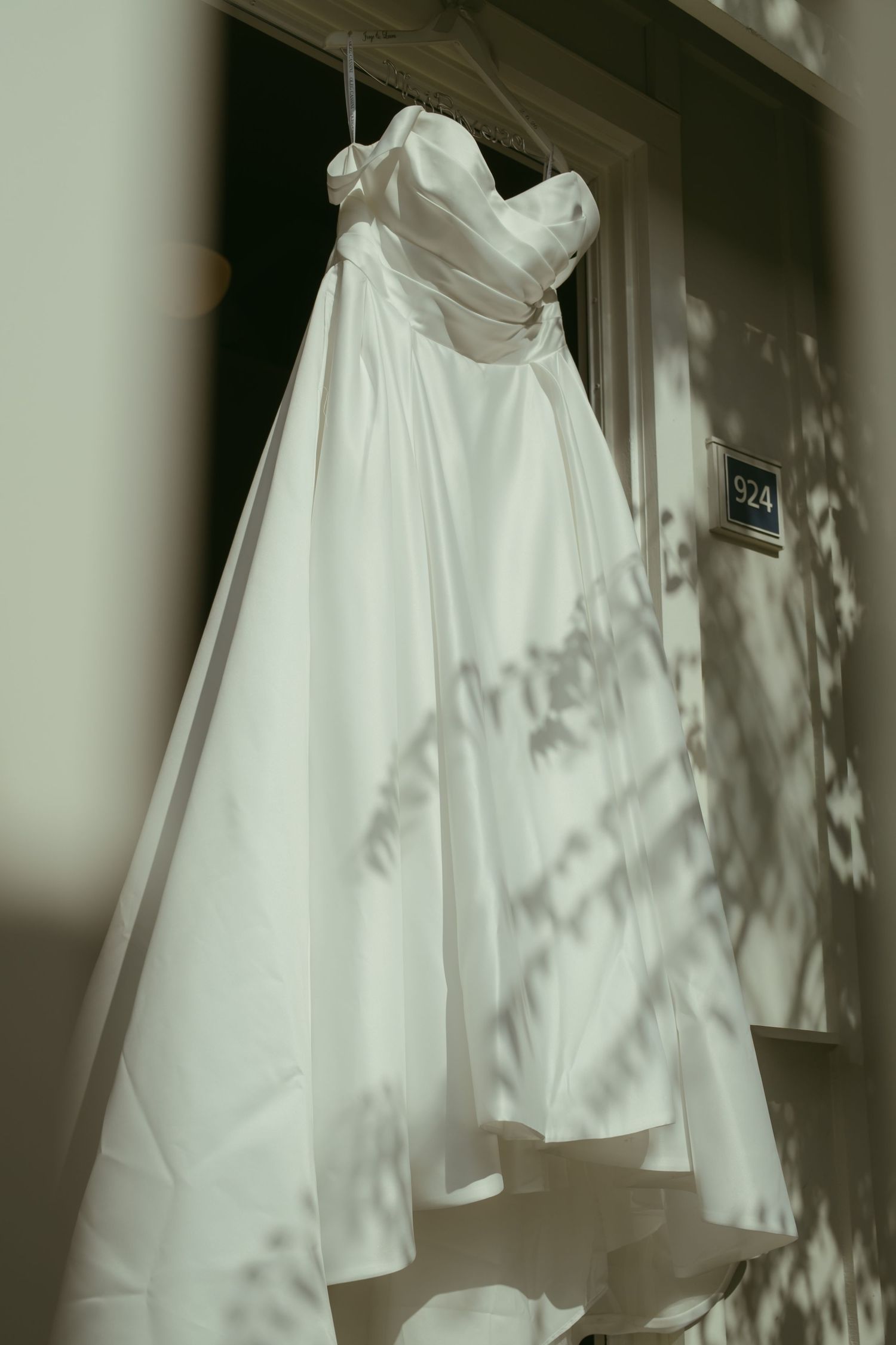White strapless satin wedding dress hanging on a door frame with dappled sunlight shadows.