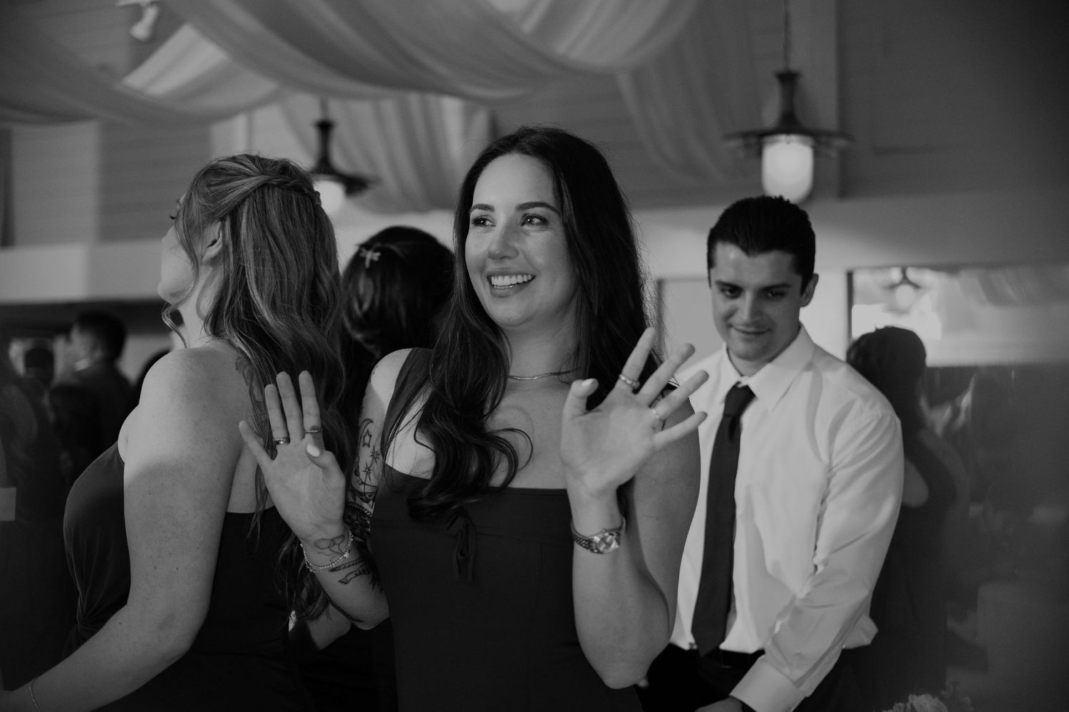 Black and white photo of a smiling woman waving at a wedding reception with guests in background.