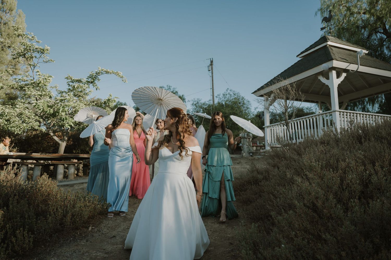 A bride in white gown walks with bridesmaids in colorful dresses holding parasols near a gazebo.