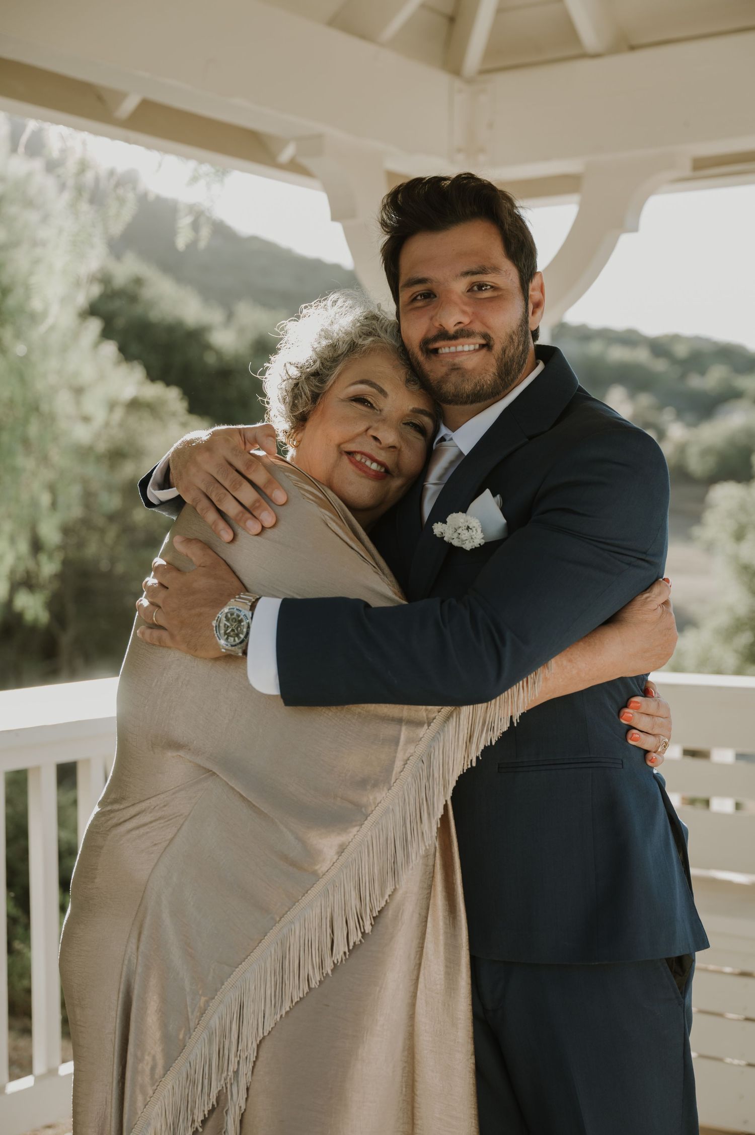 Groom in navy suit embracing older woman in gold shawl under white gazebo.