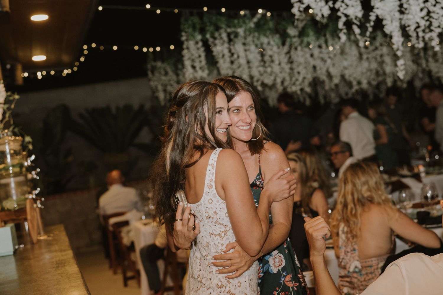 Two women share a warm embrace at an outdoor wedding reception with string lights and floral decor.