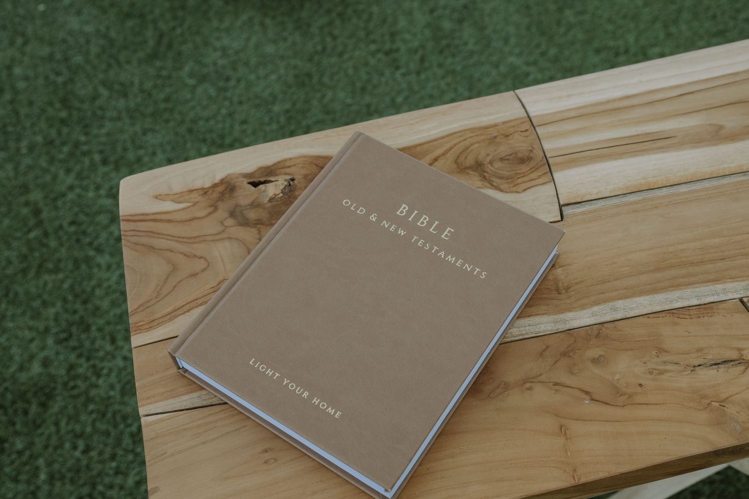 A tan hardcover Bible with Old and New Testaments resting on a wooden bench outdoors.