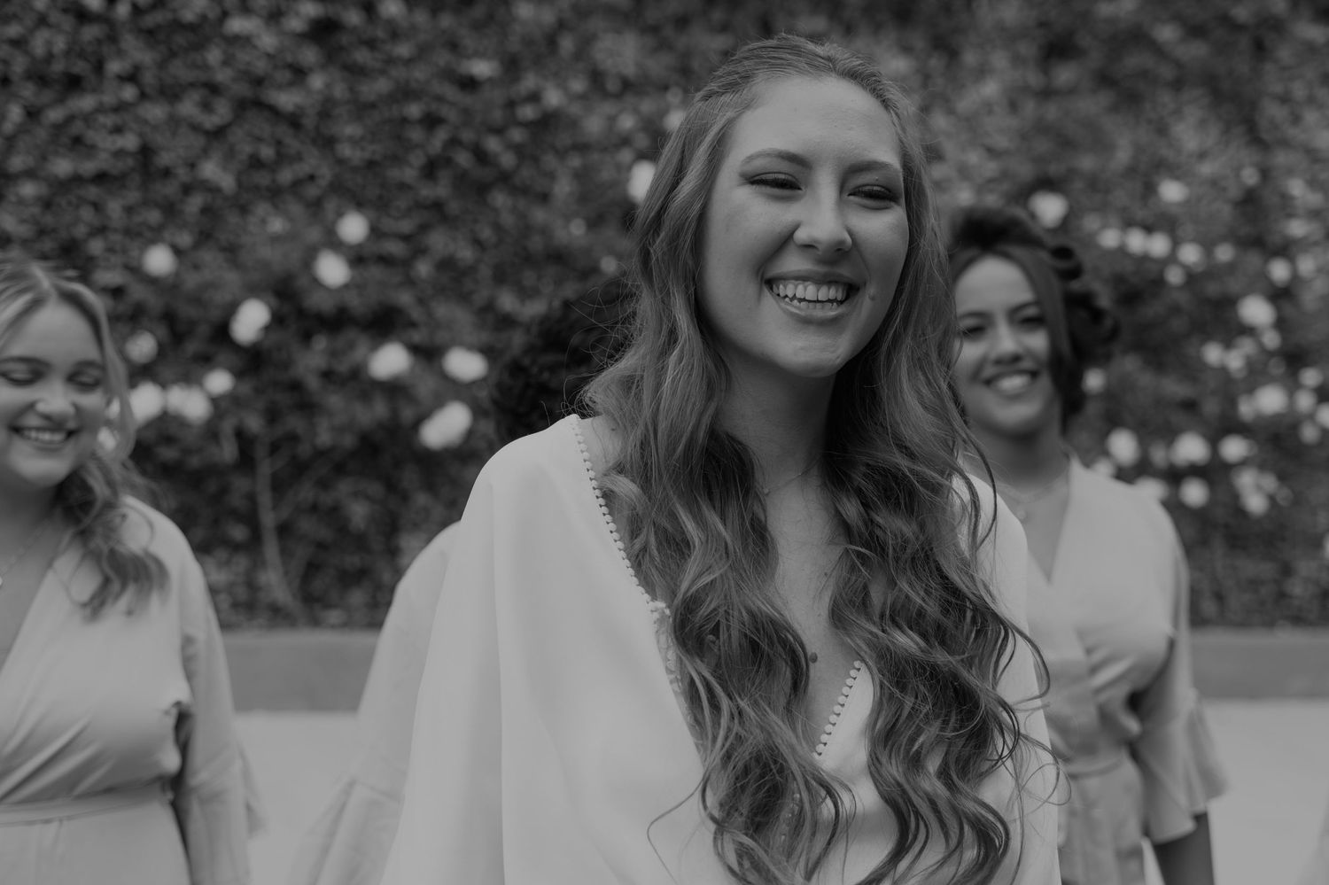 Smiling bride with long wavy hair in black and white photo with bridesmaids at outdoor wedding.