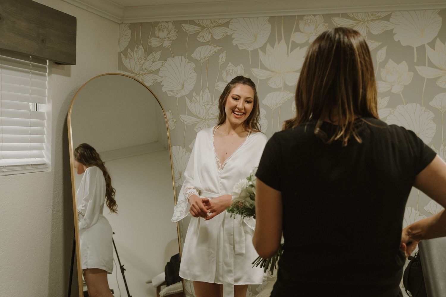 Bride in white robe smiling at reflection in arched mirror while assistant helps with preparations.