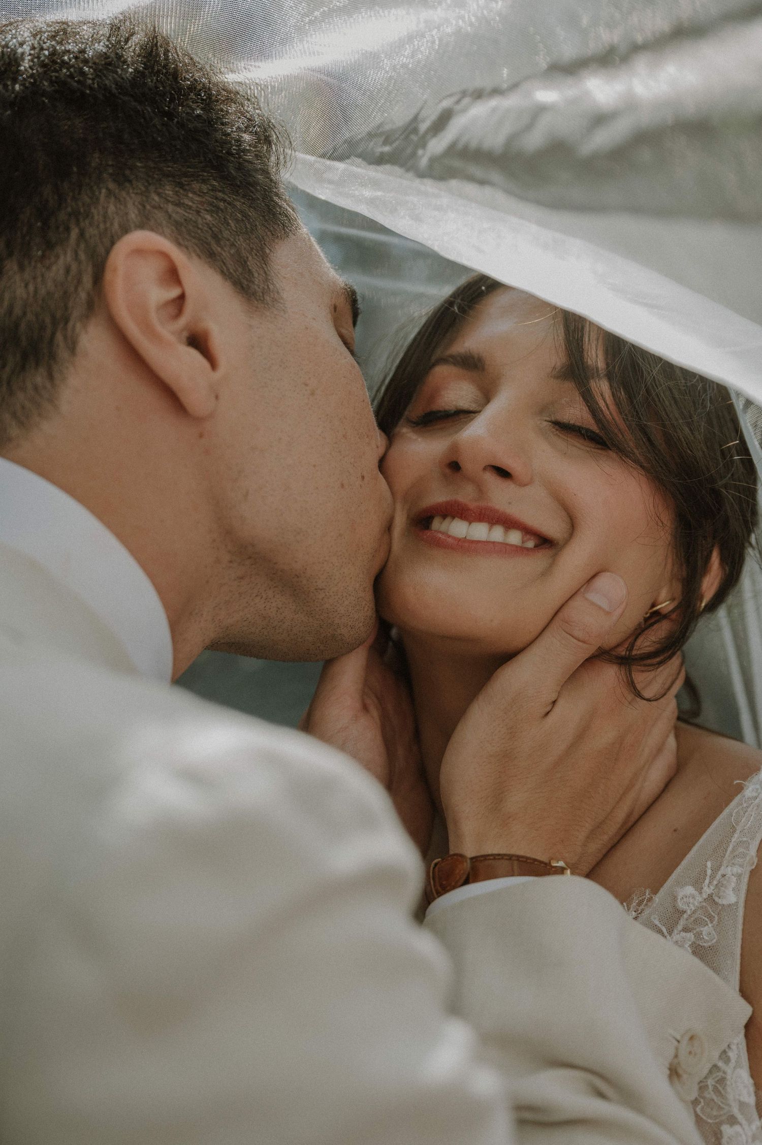Groom tenderly kisses smiling bride on the cheek under a wedding veil.