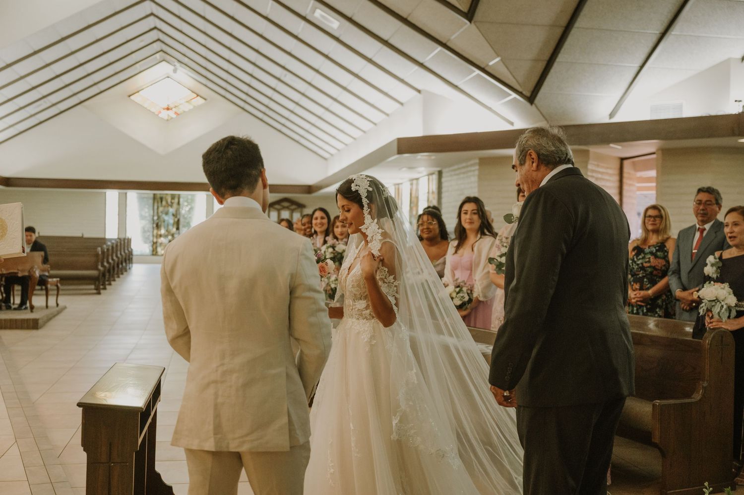 Bride and groom exchange vows at the altar surrounded by bridesmaids in a sunlit wedding chapel.
