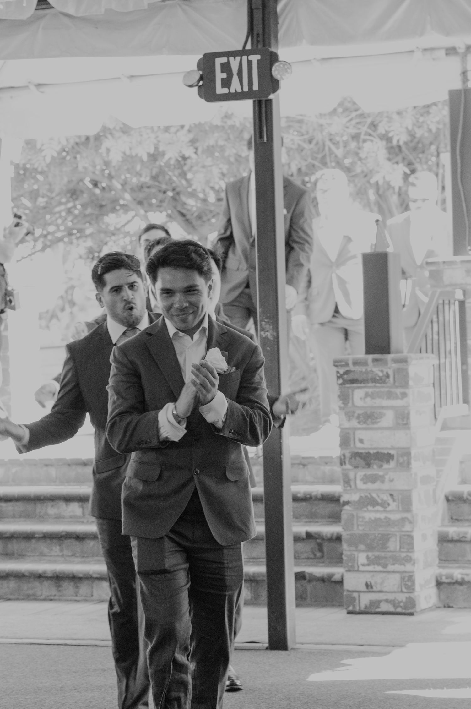 Groomsmen in gray suits walking at a wedding ceremony near an exit sign in black and white photo.