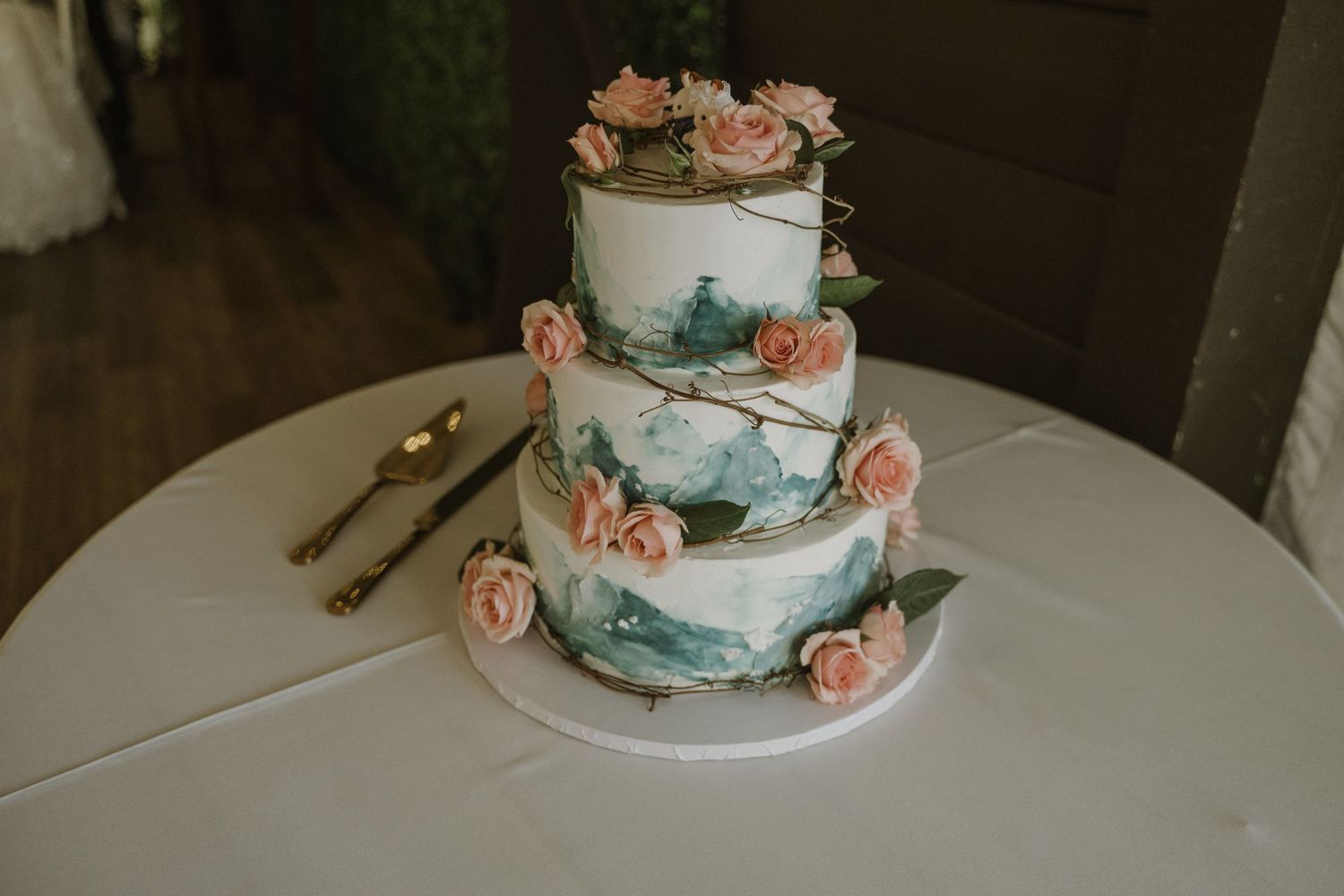 Three-tier white wedding cake decorated with pink roses and teal watercolor brushstrokes on a white tablecloth.