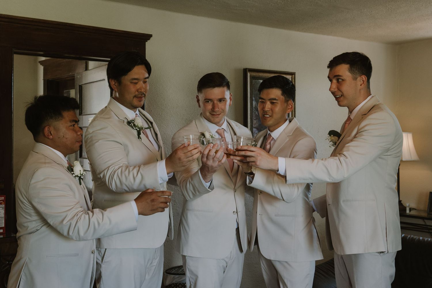 Five groomsmen in beige suits admire wedding rings together in a bright room before the ceremony.