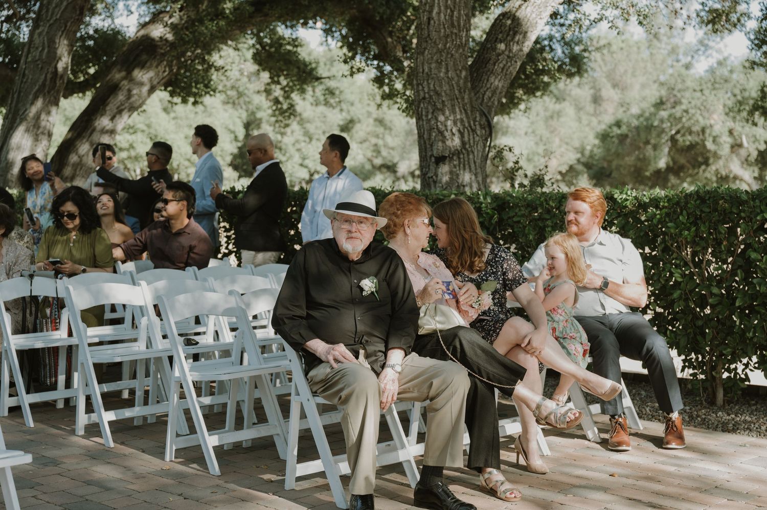 Wedding guests seated on white chairs under large oak trees at outdoor ceremony venue.