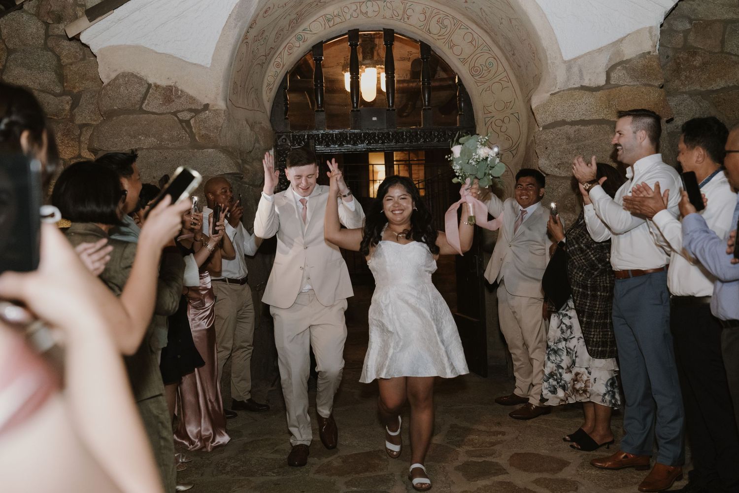 Newlyweds joyfully exit through a stone archway as guests cheer and applaud at their wedding.
