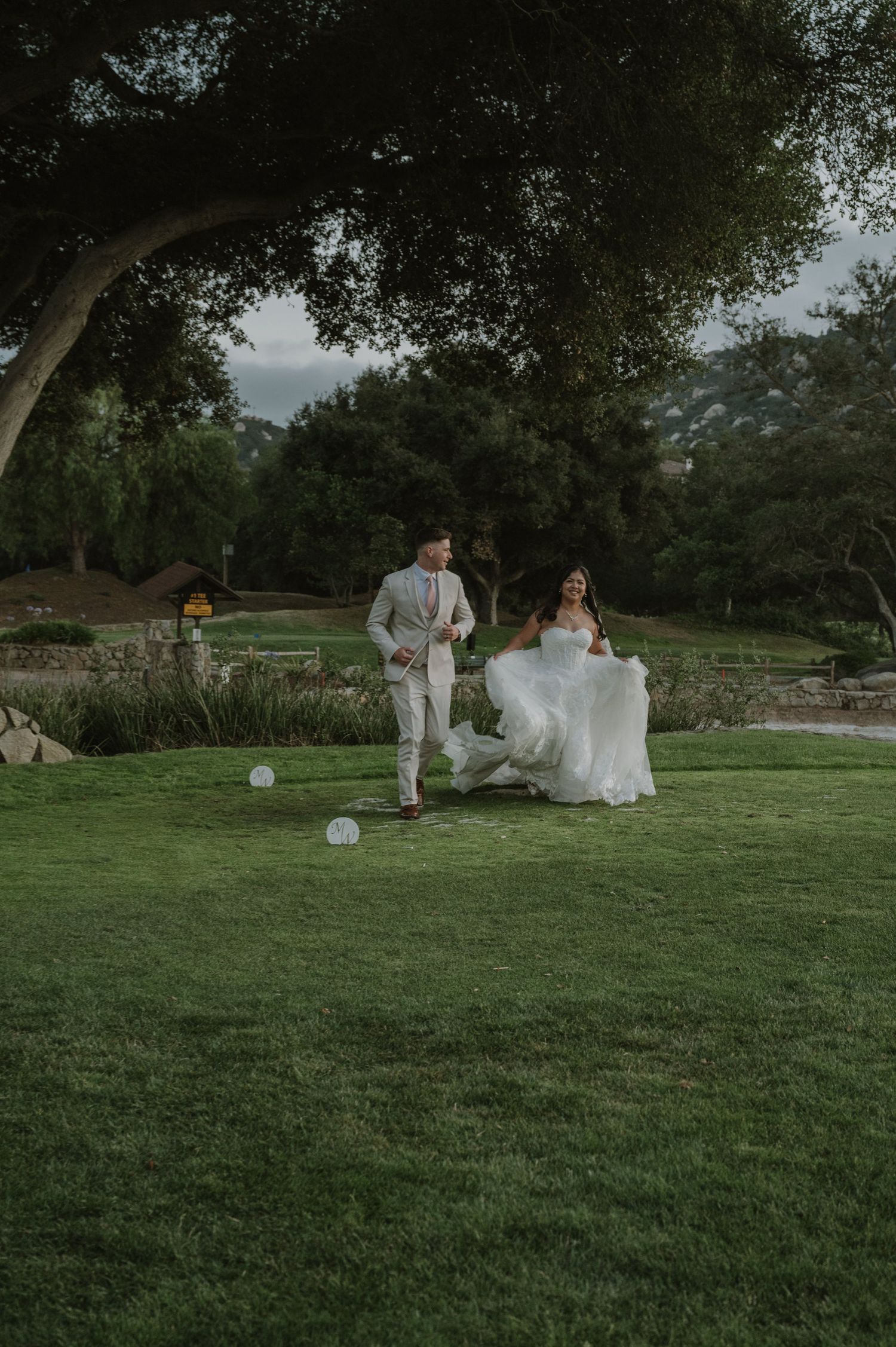 Bride and groom walking on lush green lawn under large oak tree at outdoor wedding venue.