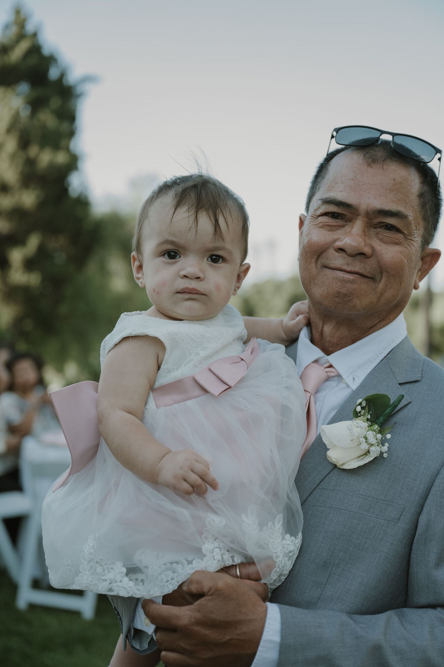 Older man in gray suit with boutonniere holding a baby girl in white and pink dress at a wedding.