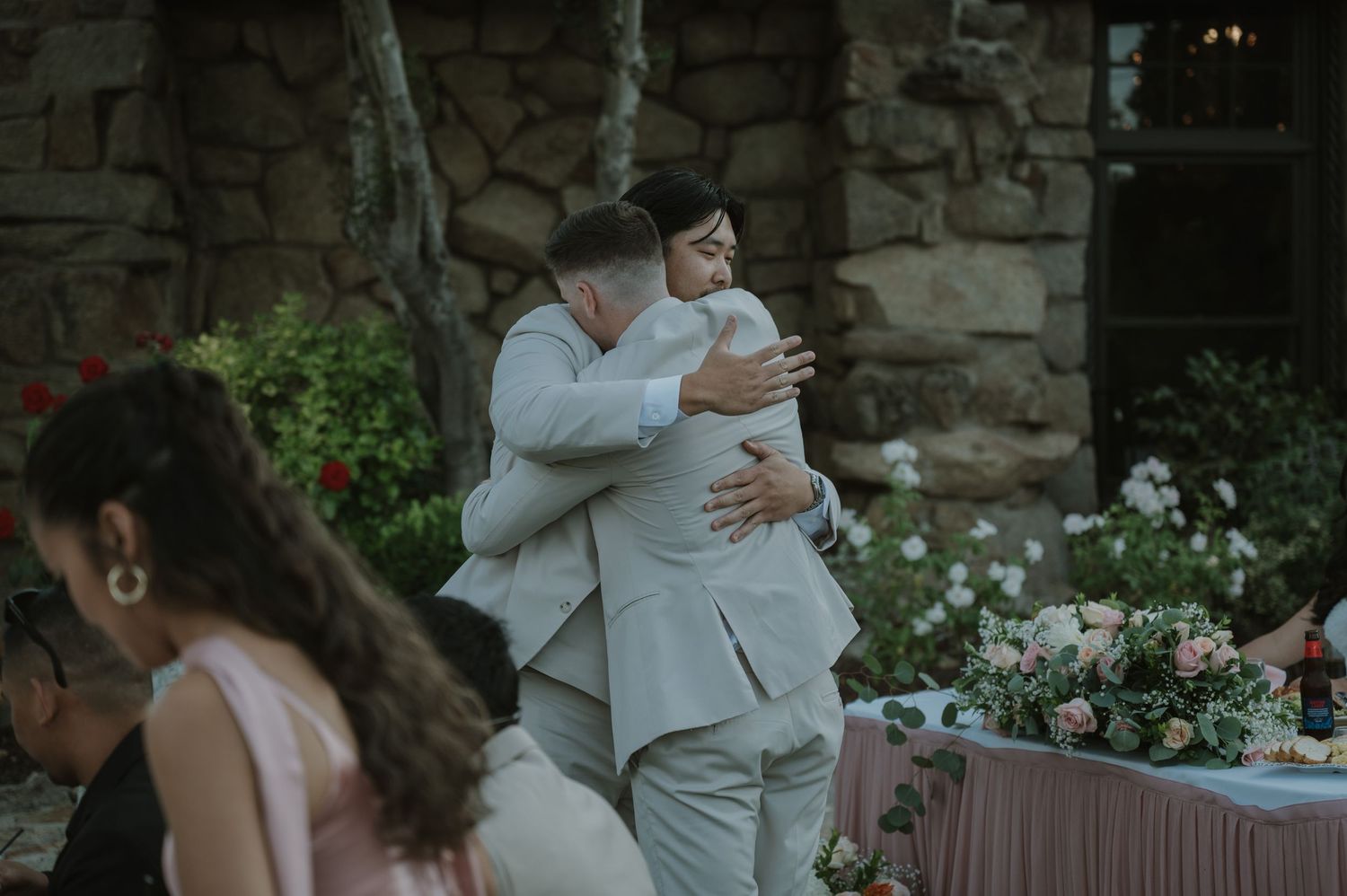 Two grooms share a heartfelt embrace at their outdoor wedding reception near a stone building.