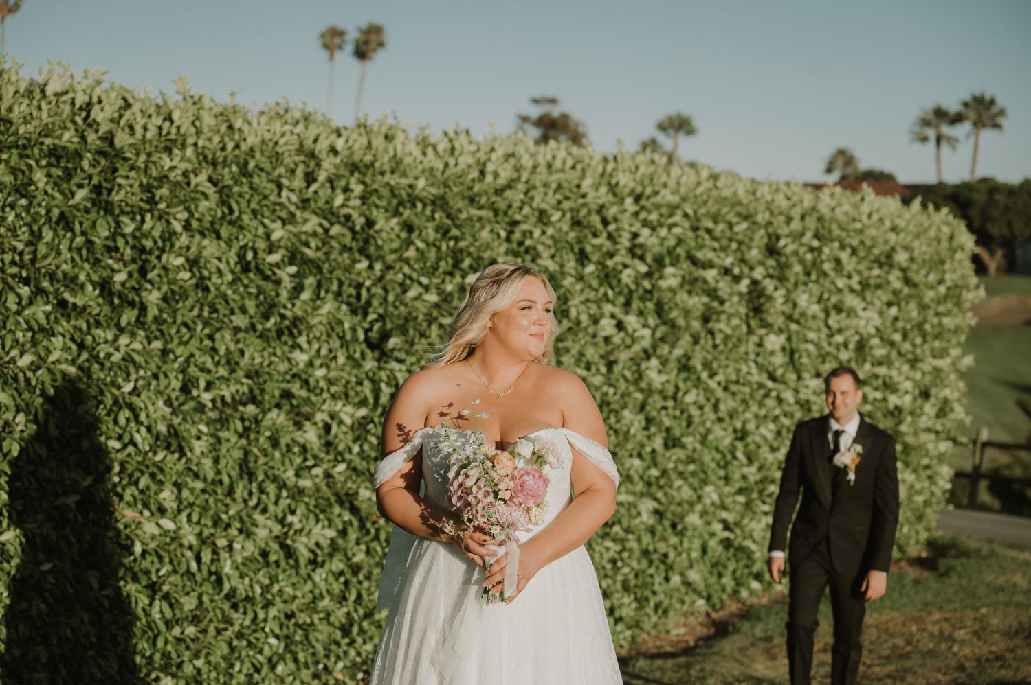 Blonde bride in off-shoulder gown holds heart-shaped bouquet near lush hedge as groom approaches in tuxedo.