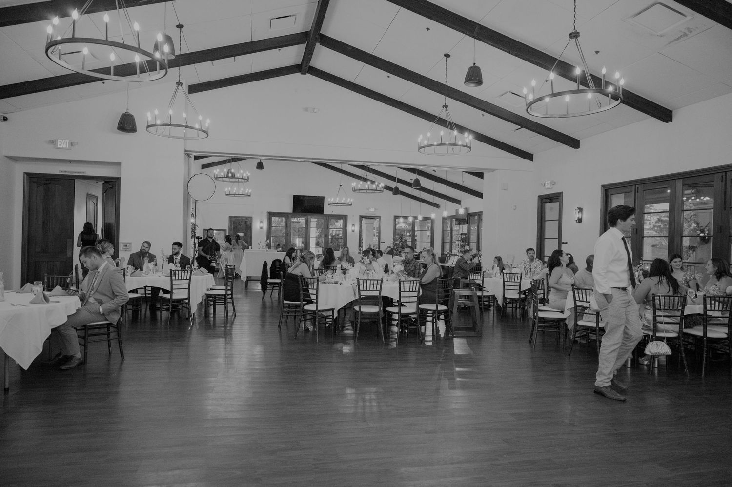 Elegant wedding reception hall with vaulted ceilings, chandeliers, and guests seated at round tables in black and white.