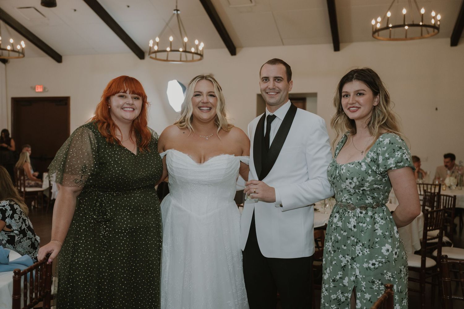 Bride and groom posing with two female wedding guests in a decorated reception venue with chandeliers.