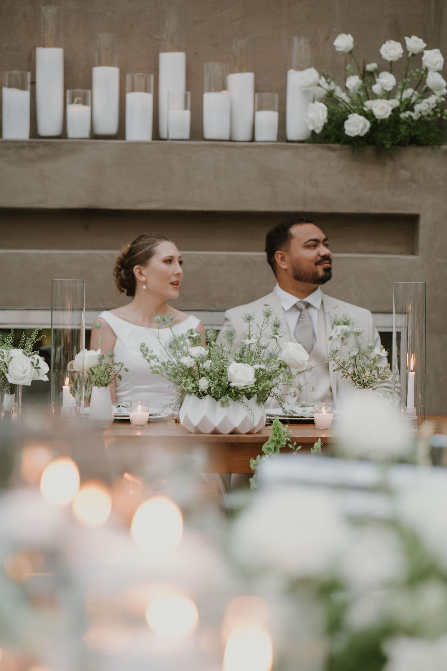 Bride and groom seated at elegant candlelit sweetheart table with white floral arrangements at their wedding reception.