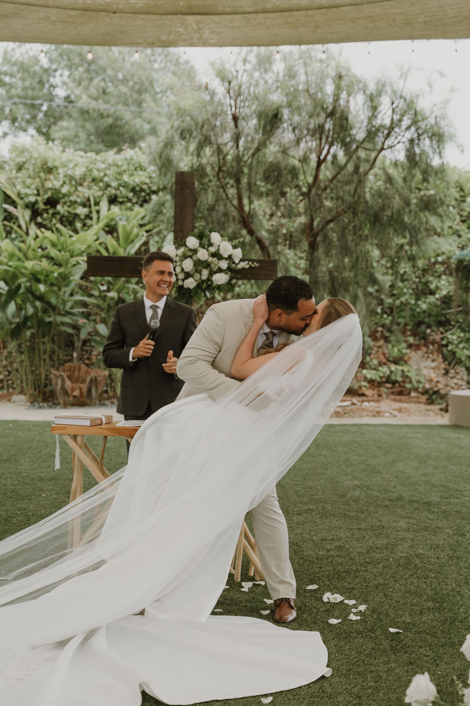 Groom dips bride for a kiss at outdoor wedding ceremony while officiant smiles in background.