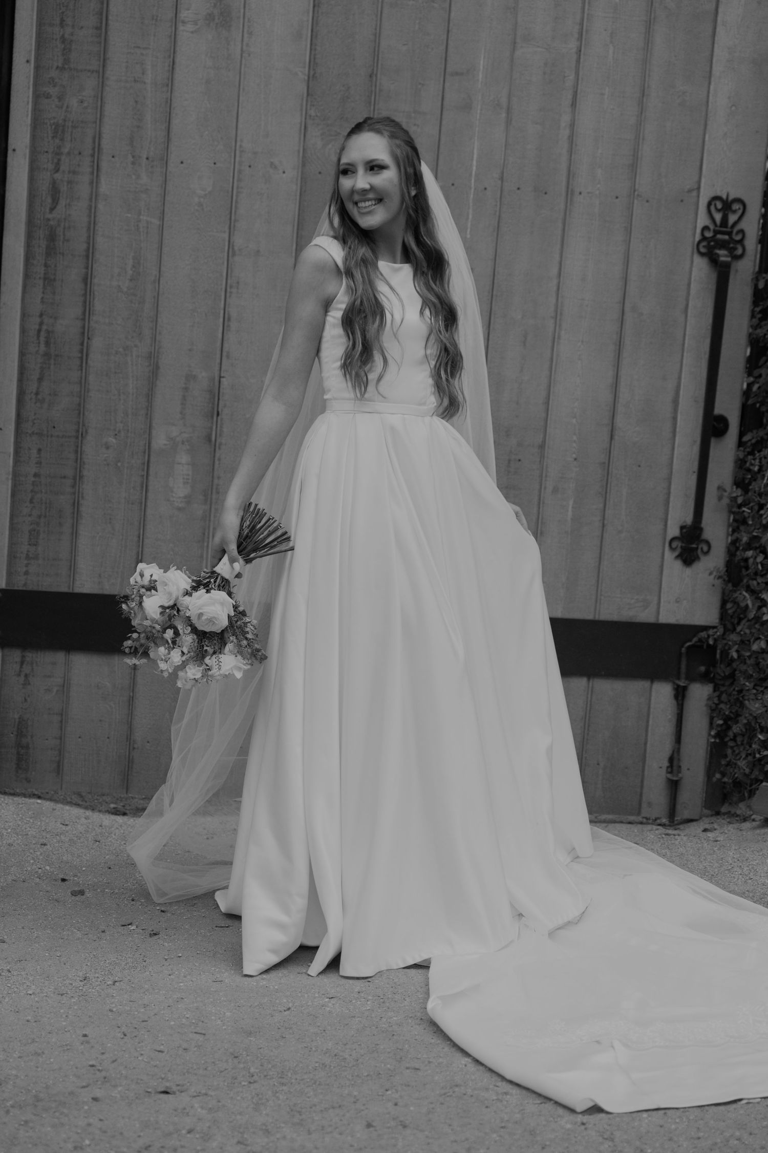 Elegant bride in white sleeveless gown holding floral bouquet posing before rustic barn doors.