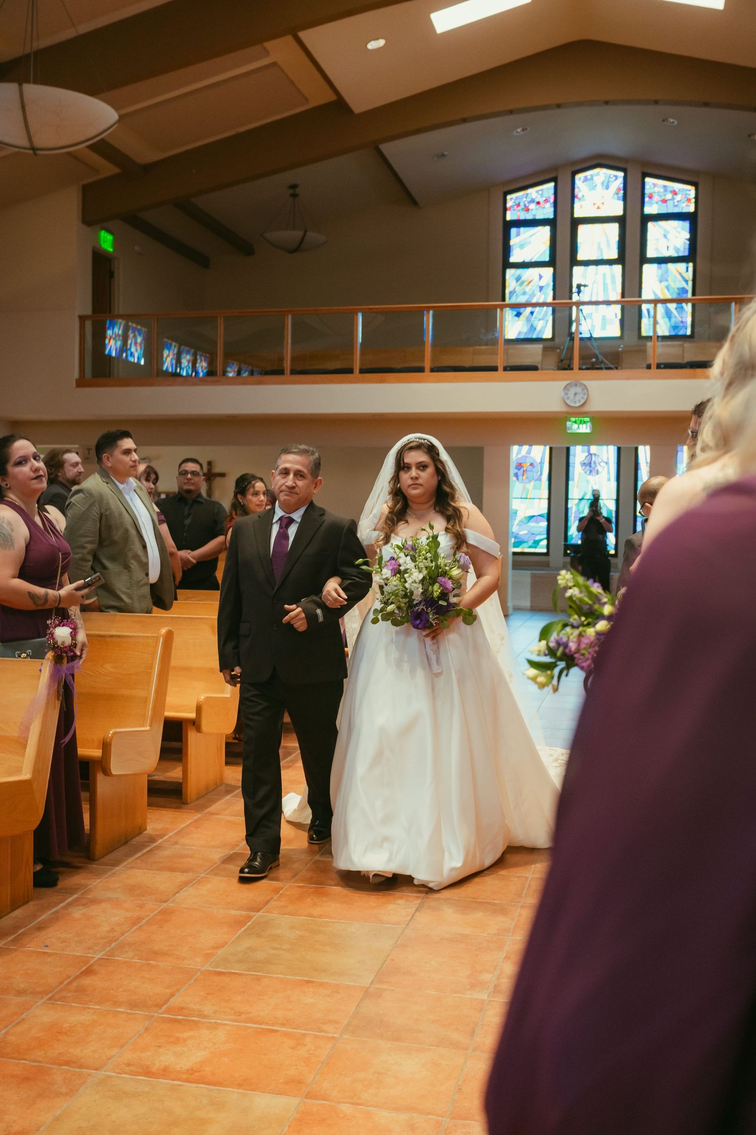 Bride in white gown walks down church aisle with escort, holding purple bouquet, stained glass windows in background.