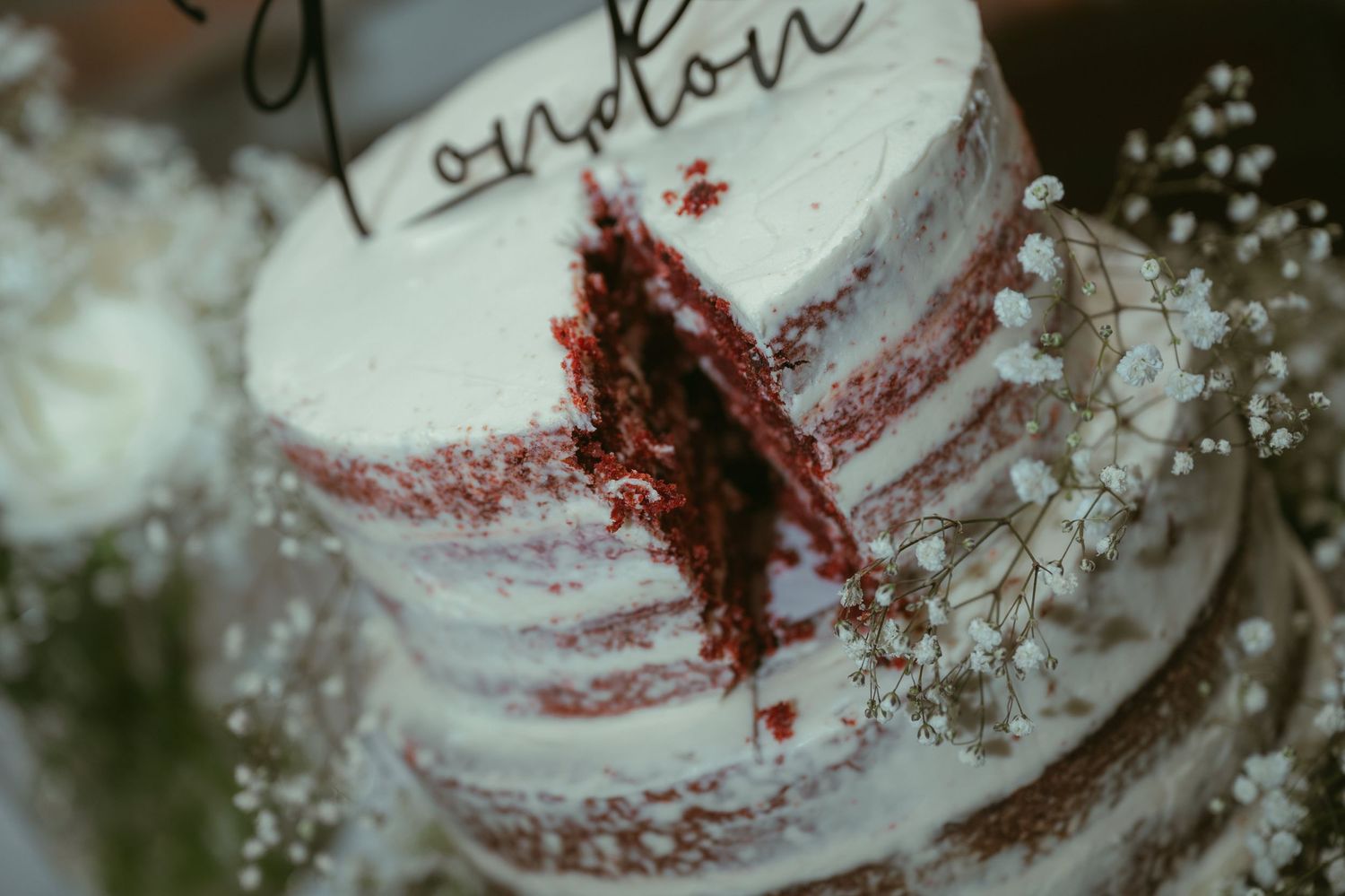 A sliced red velvet naked wedding cake with white frosting and a London cake topper, surrounded by baby's breath flowers.