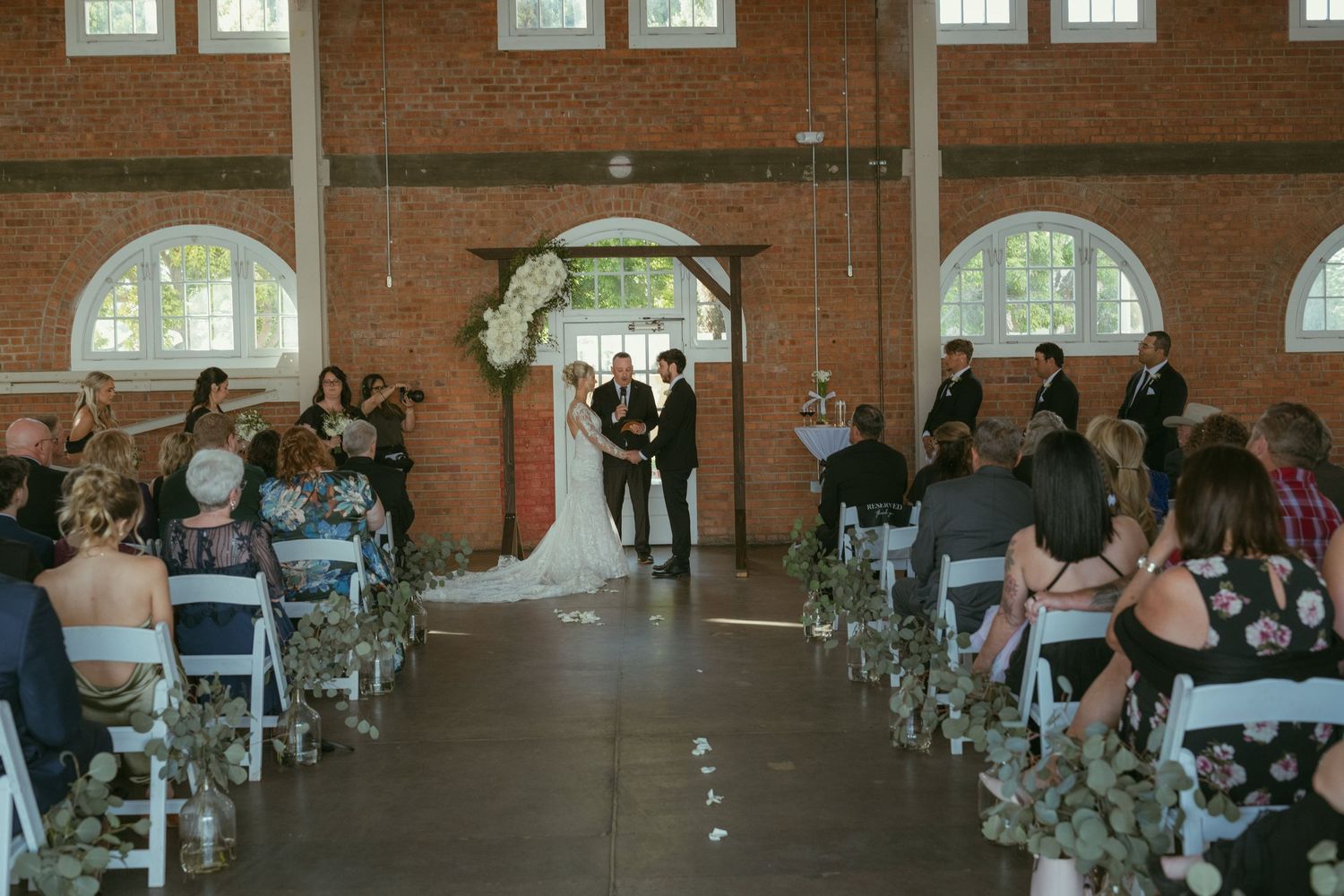 Elegant indoor wedding ceremony in brick venue with floral arch, aisle decorations, and seated guests in white chairs.