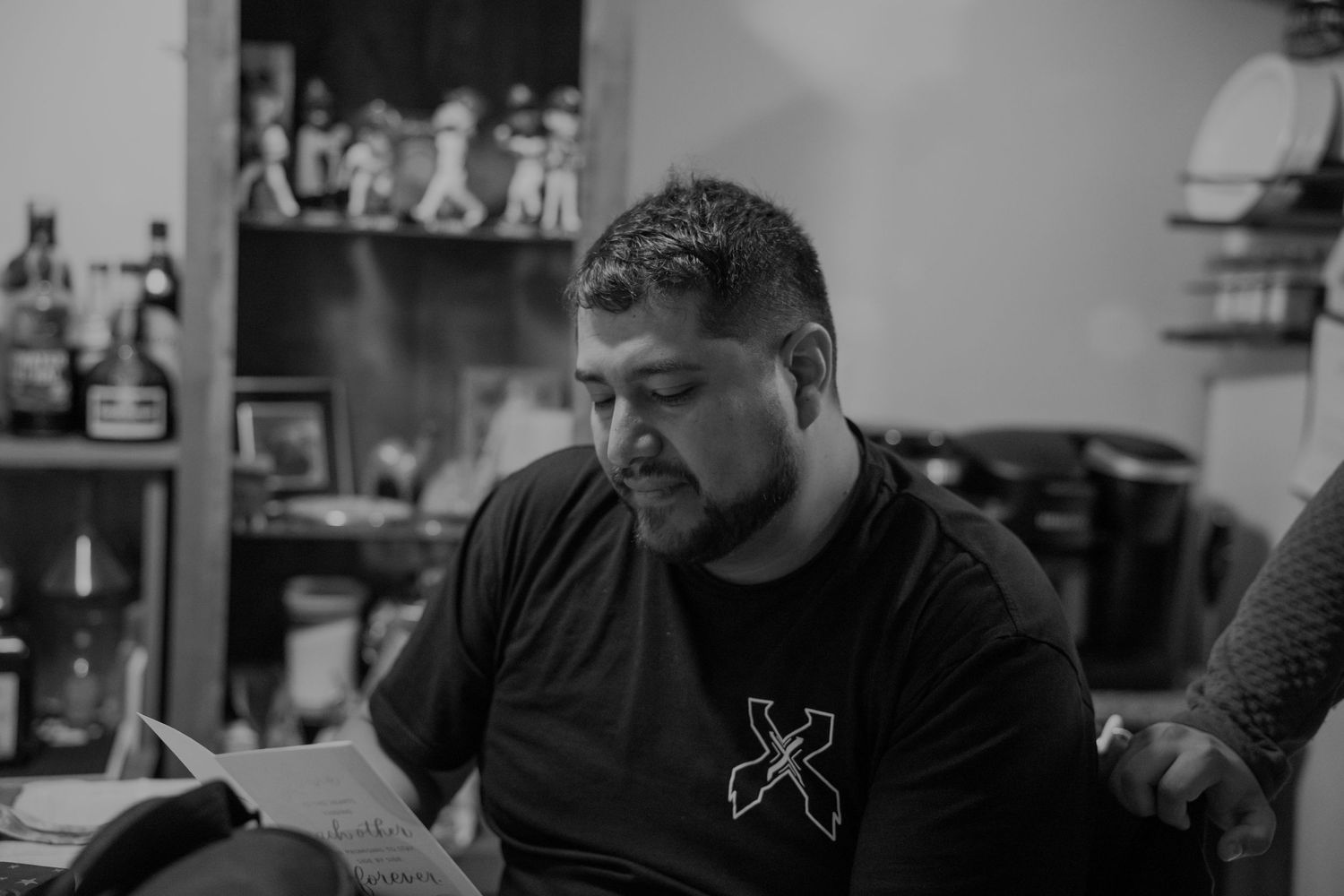 Black and white photo of a man in a black t-shirt reading a document indoors with shelves in the background.